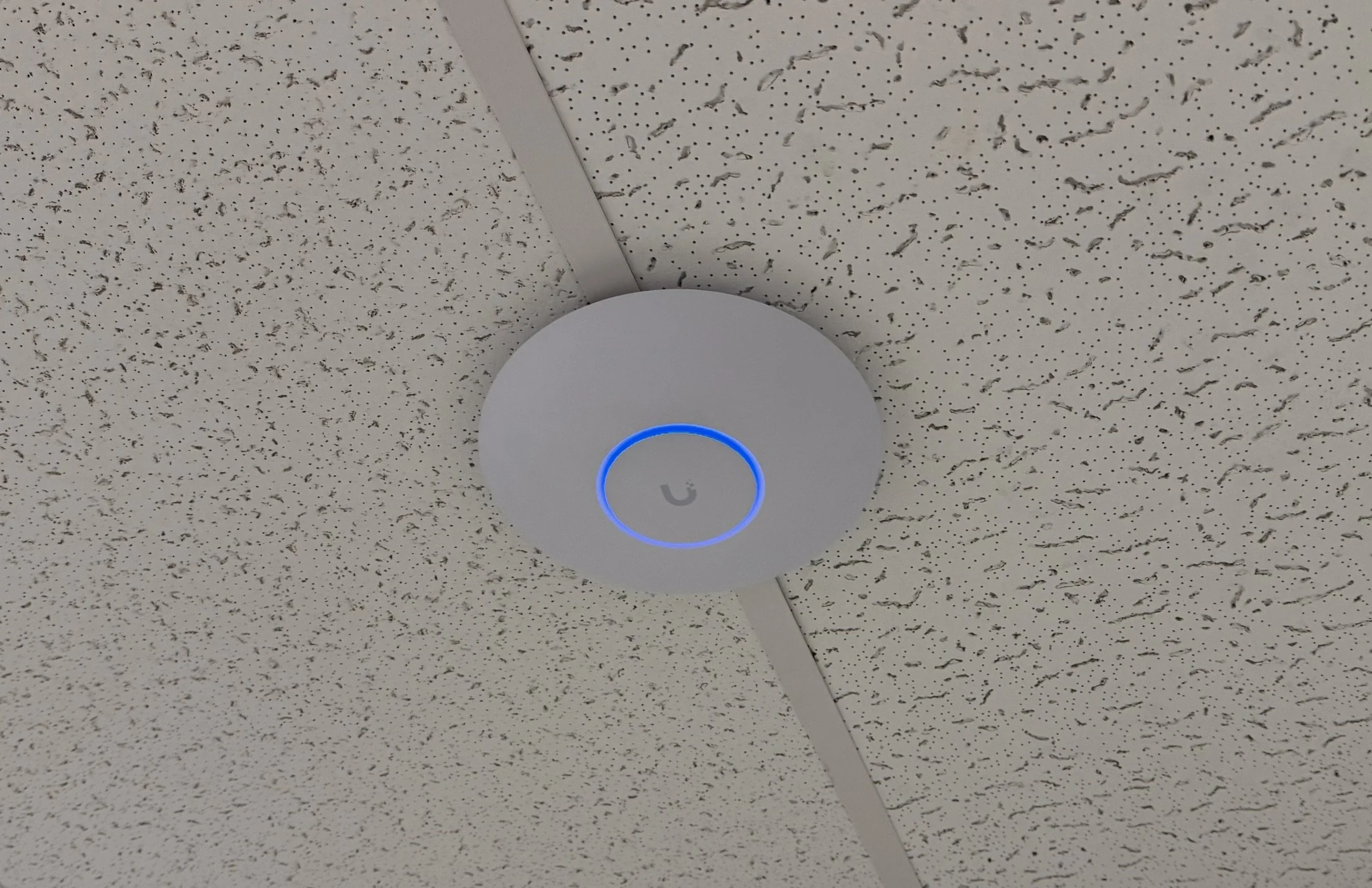 Ceiling-mounted device with a circular blue-lit indicator and a symbol in the center, on a textured ceiling.