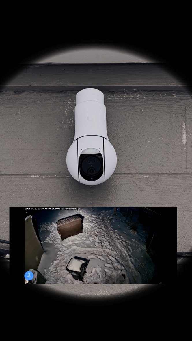 Top of a white security camera mounted on a gray wooden wall, with a split view showing a snowy backyard with a trash bin, a fence, and a snow-covered pathway.