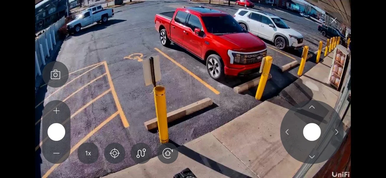 A parking lot with several parked vehicles, including a prominent red pickup truck in the center, surrounded by yellow safety bollards and parking space markings.