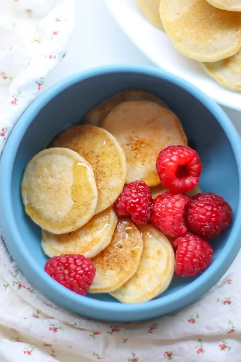 A blue bowl filled with small pancakes topped with syrup and fresh raspberries, with additional pancakes on a white plate nearby.