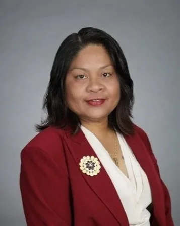 A woman with shoulder-length black hair wearing a red blazer, a white blouse, and a floral brooch, posing against a gray background.