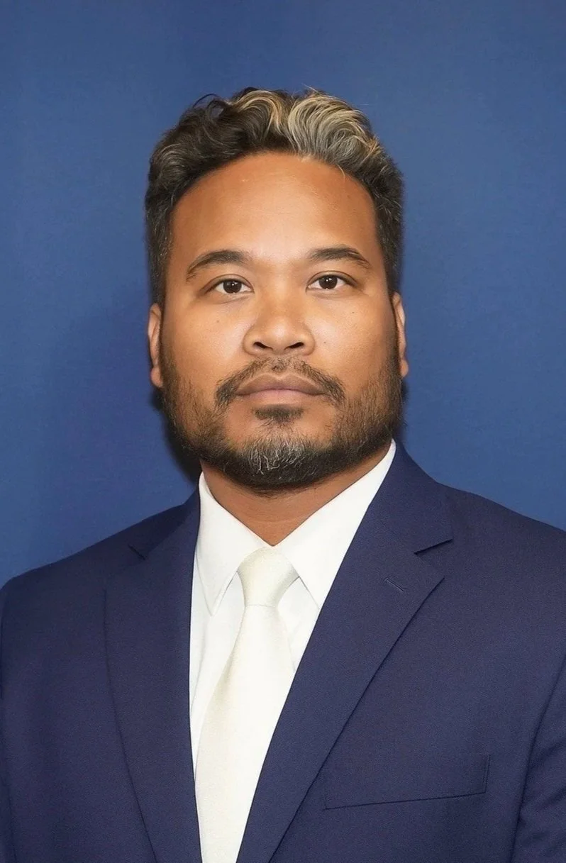 A man in a navy blue suit, white shirt, and white tie, with short dark hair and a beard, is posed against a plain blue background.