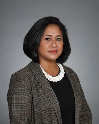 A woman with shoulder-length black hair wearing a brown plaid blazer, a black top, and a pearl necklace, standing against a plain gray background.