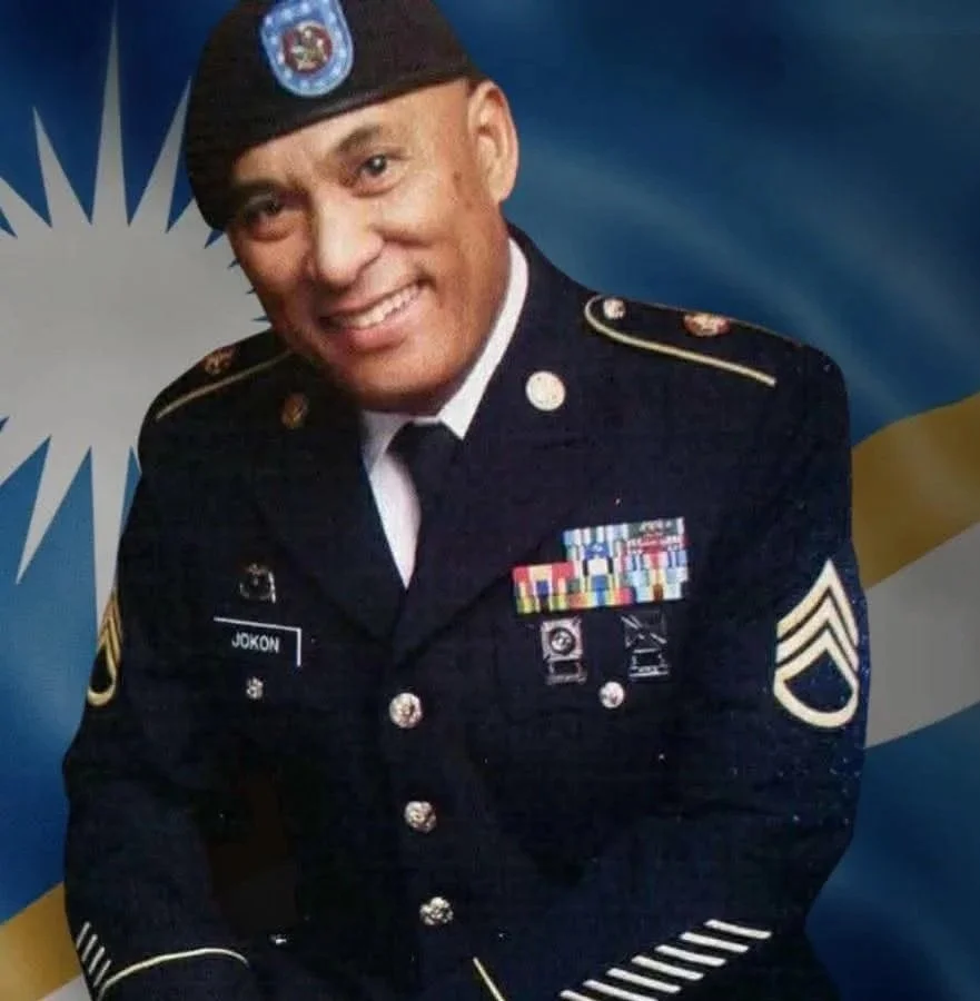 A smiling man in a military uniform with medals and patches, wearing a beret, against a background featuring the flag of Ukraine.