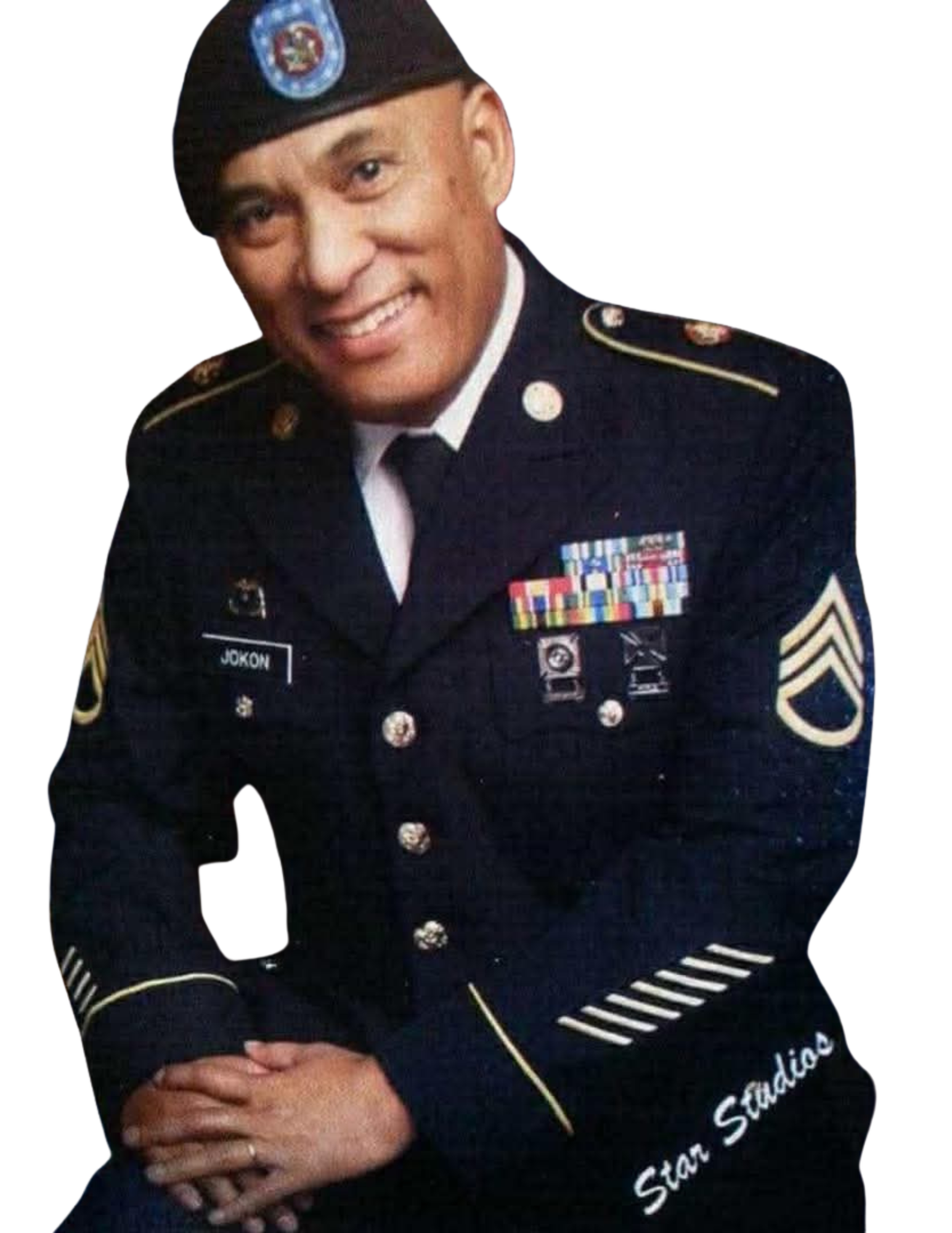 A smiling man in a United States Army uniform with various ribbons and ranks, wearing a beret with a badge, sitting with his hands clasped.