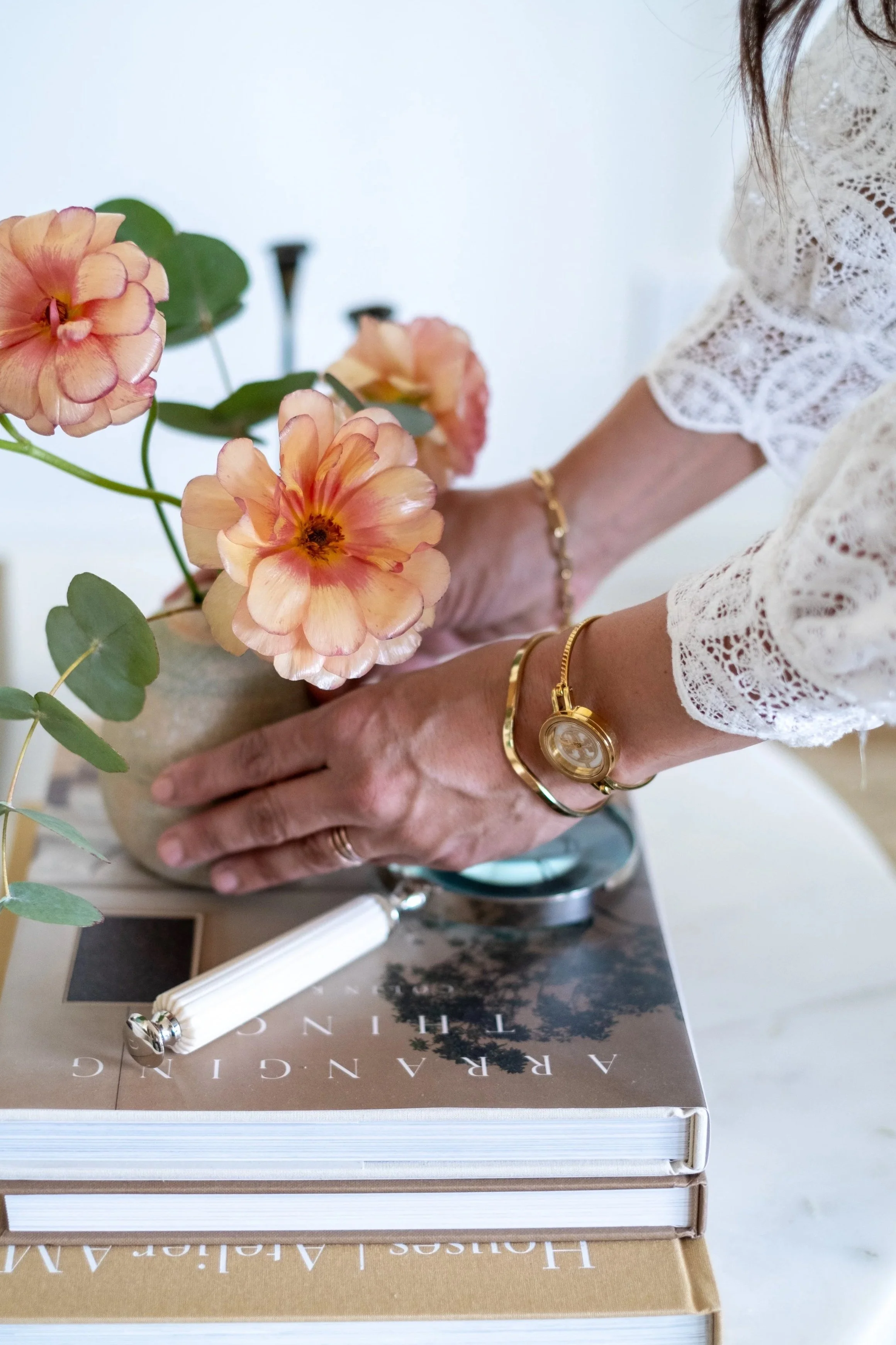 A person arranging pink or peach-colored flowers in a beige vase on a white table. The person is wearing a white lace top and gold jewelry, including a bracelet and a watch. There are two large books on the table with the open pages facing down, and a white object, possibly a pen, rests on one of the books.