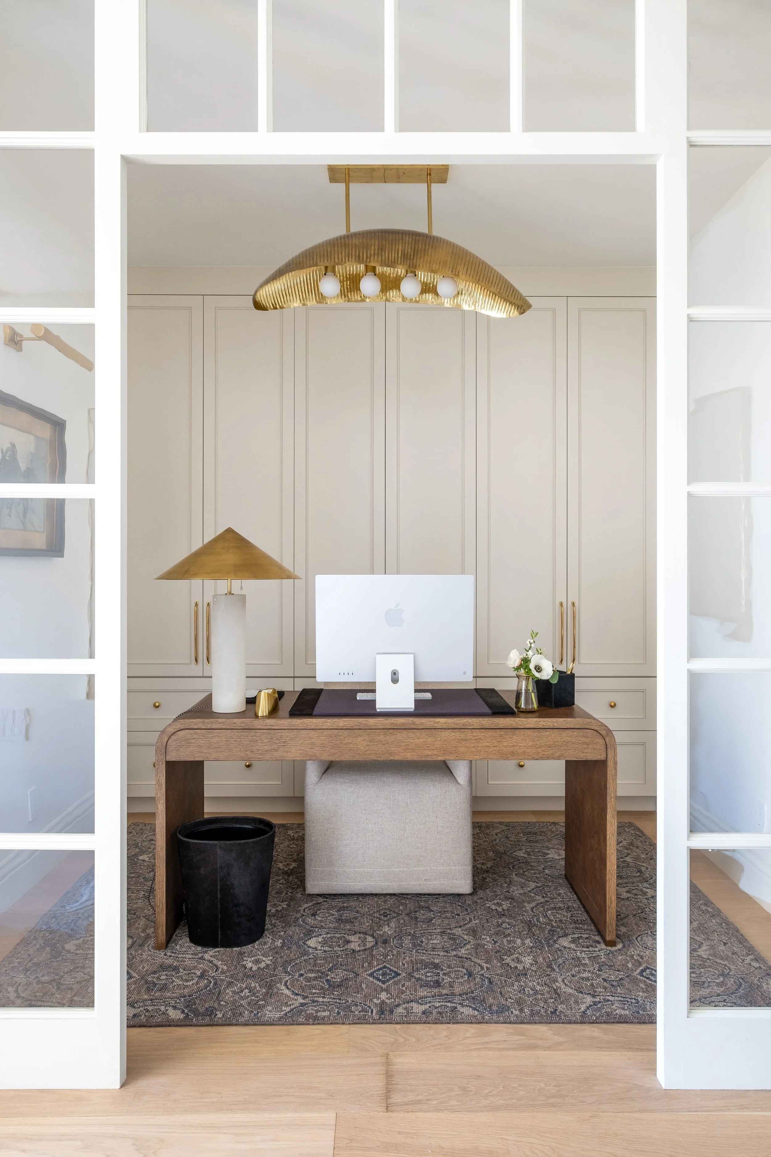 Modern home office with a wooden desk, vases with white flowers, a gold and white table lamp, an Apple computer, and a gold and white overhead light fixture.