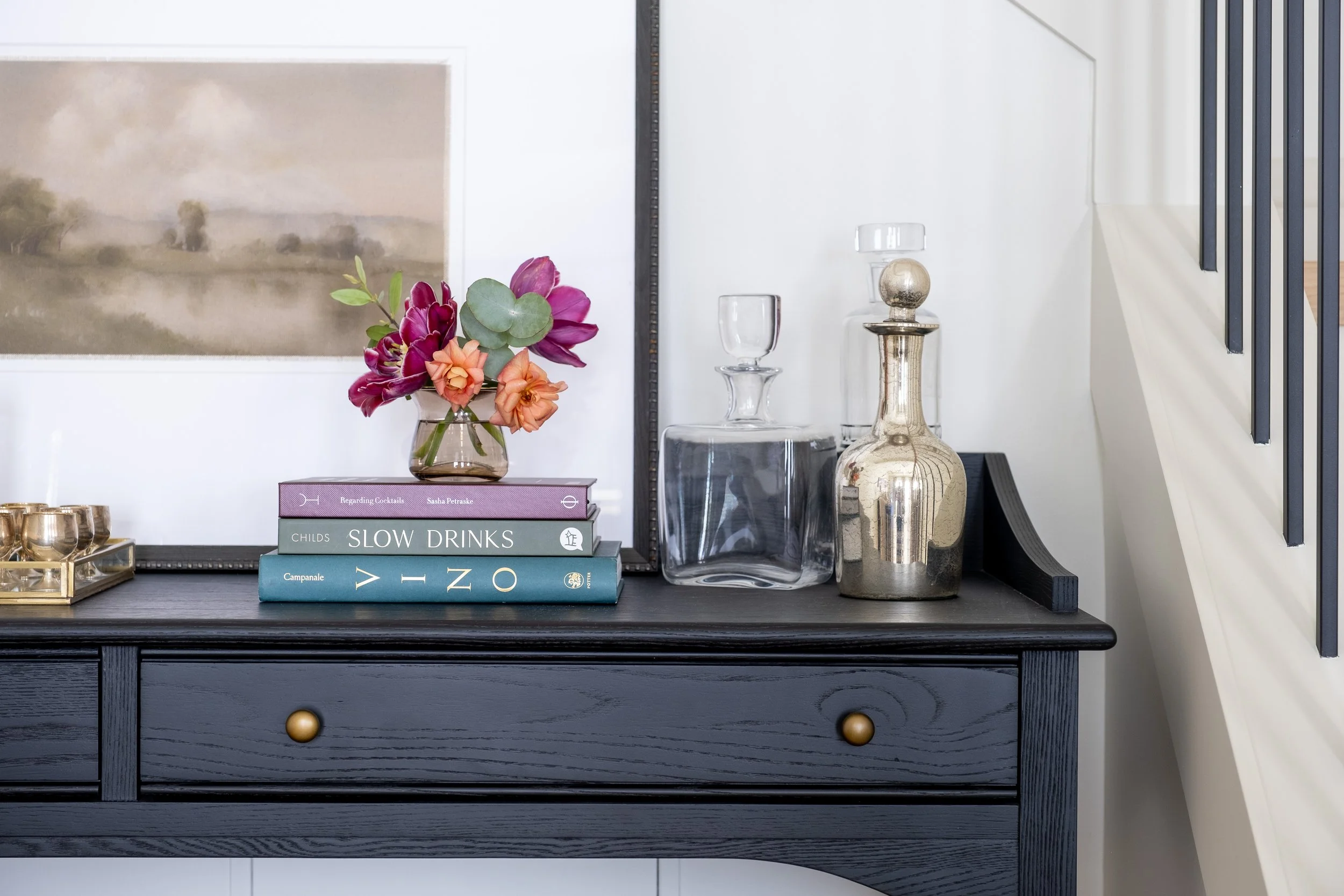 A black wooden console table with a stack of three books, a vase with pink, purple, and peach flowers, and various glass bottles, in front of a large mirror and beside a staircase.