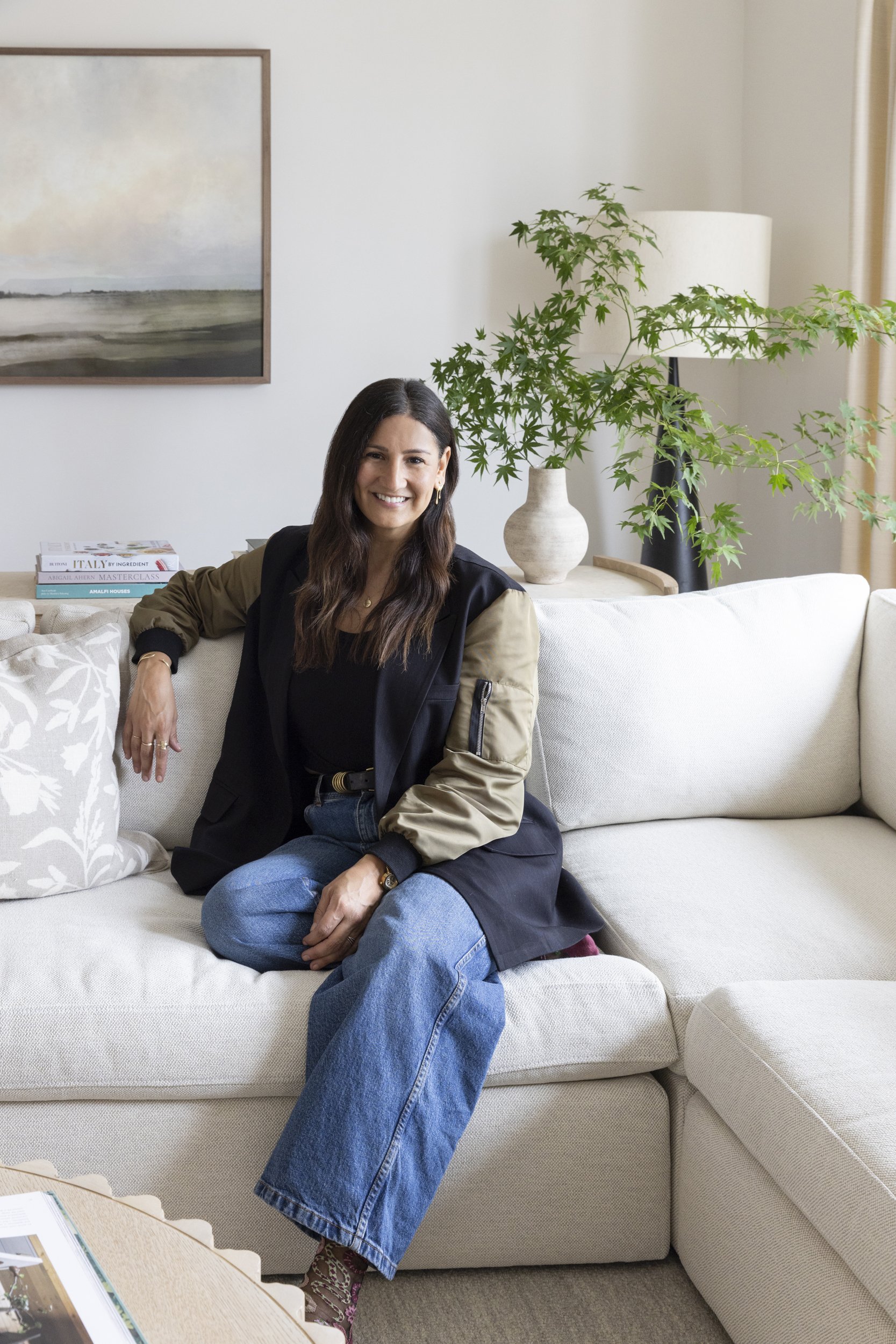 A woman with long dark hair and a warm smile sits on a cream-colored sofa in a living room. She is wearing a black top, a stylish jacket with tan sleeves, and jeans. Behind her, there is a large green potted plant, a modern floor lamp, a wooden framed painting, and some books on a side table.