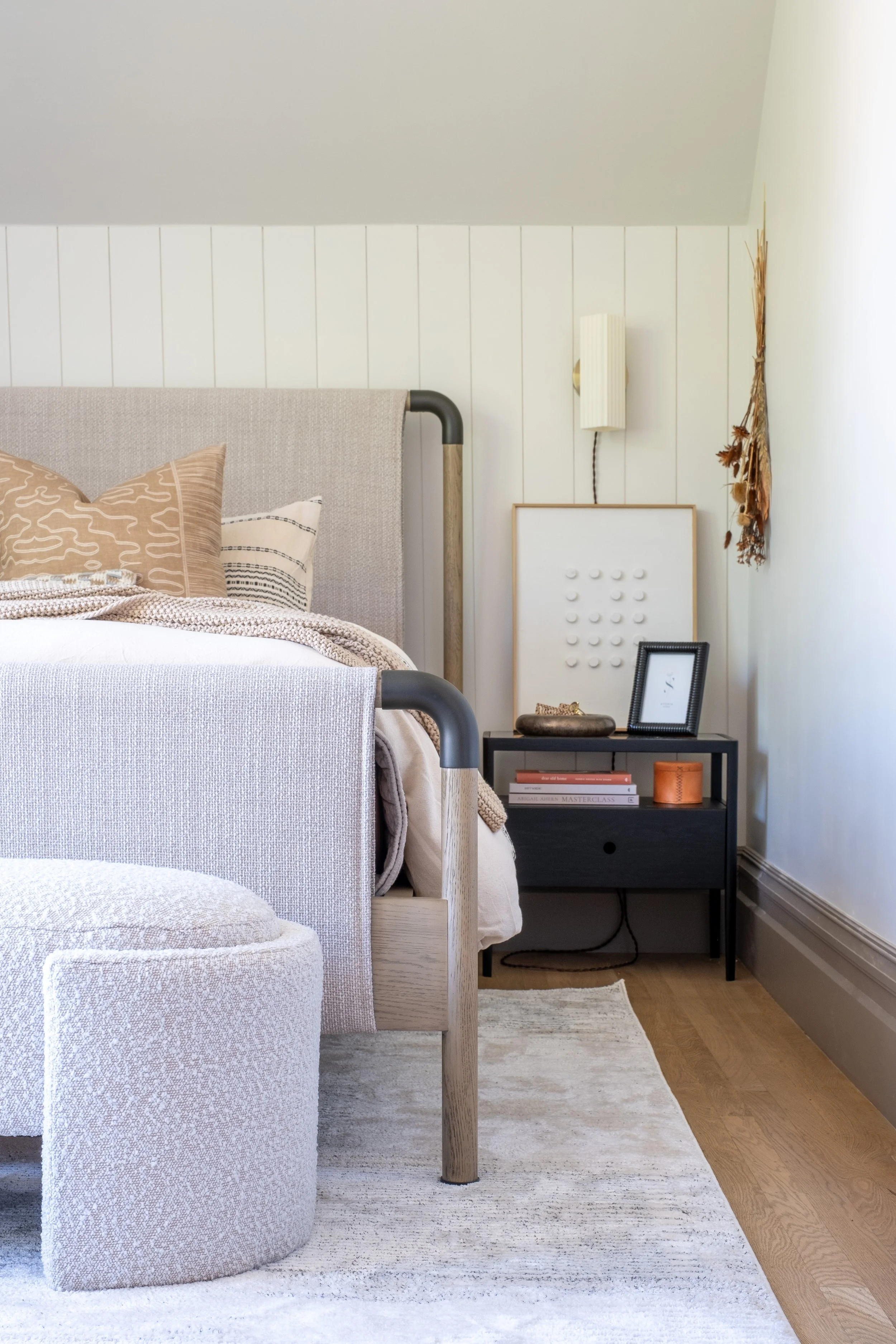 A cozy bedroom corner with a bed featuring beige and white pillows, a black nightstand with books and decorative items, and wall decor including a hanging dried plant and framed artwork.