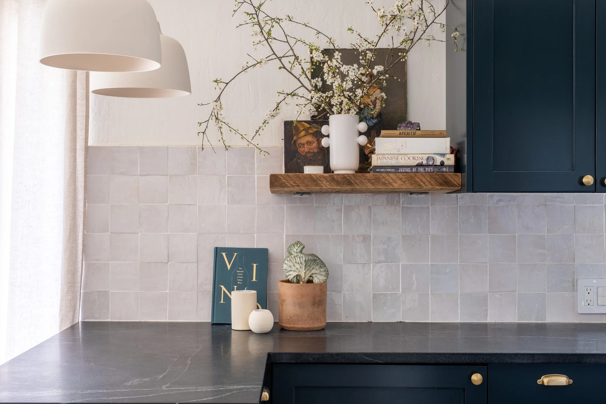 Kitchen countertop with decorative items, including potted plant, candles, picture book, and a small sculpture, against a tiled backsplash and upper cabinet.