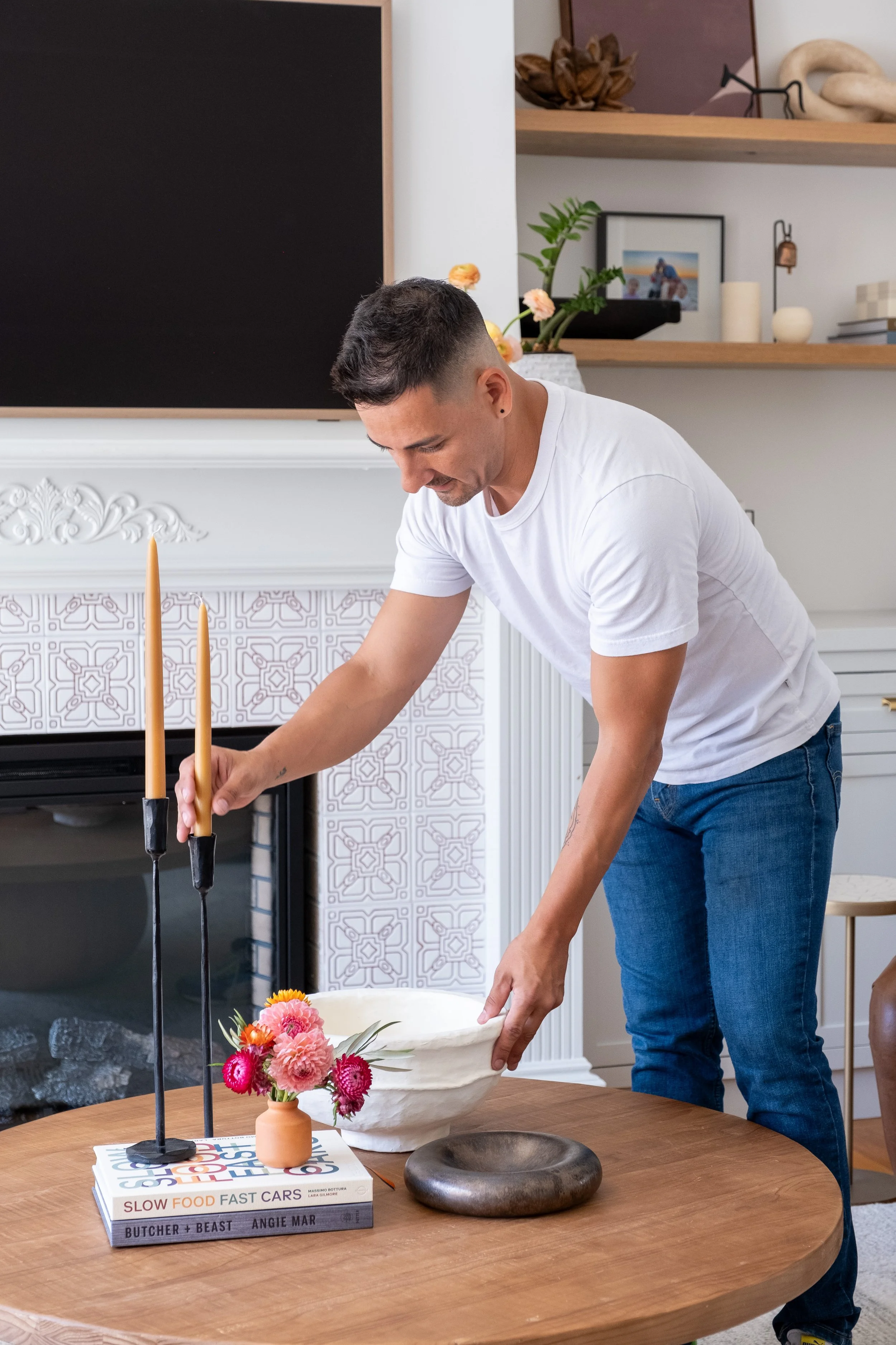 A man wearing a white t-shirt and blue jeans arranging objects on a wooden table, including a white bowl, a small vase with pink and red flowers, a black stone dish, and a stack of books, in a decorated living room.