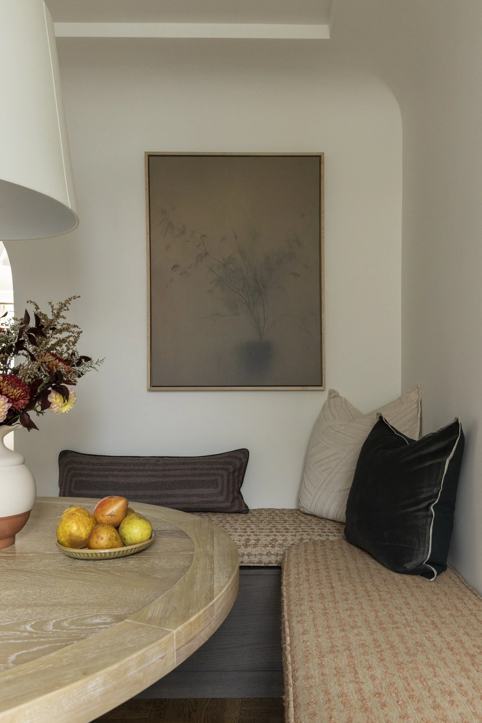 A cozy corner with a round wooden table holding a bowl of pears and apples, a large flower vase with dried flowers, and a built-in bench with multiple pillows and cushions against the wall, with a framed artwork of a vase with foliage hanging above.