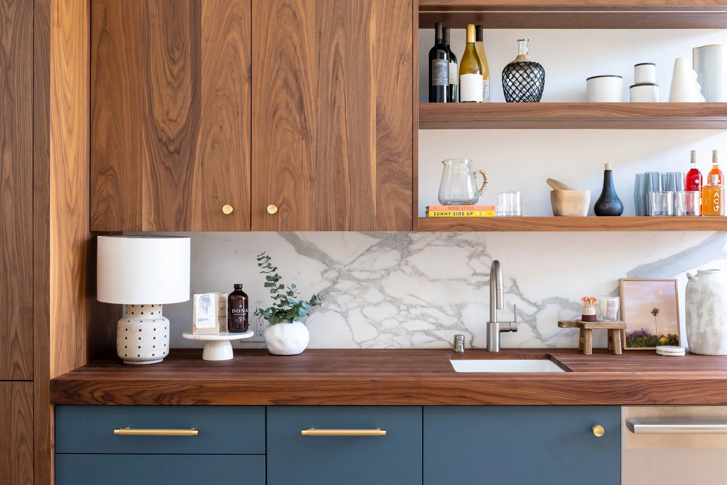 Kitchen with wooden cabinets, open shelves with decorative items and bottles, marble backsplash, blue lower cabinets, and a wooden countertop with a small sink.
