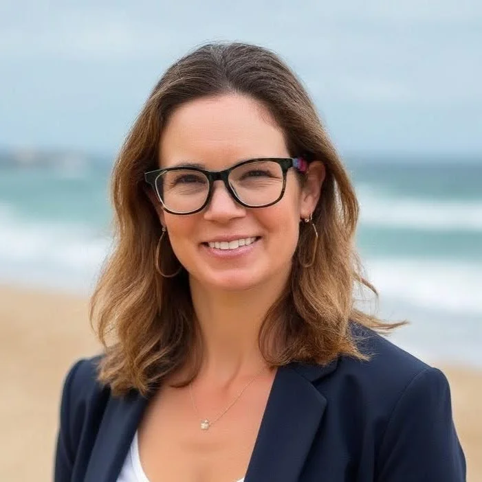 A woman with shoulder-length brown hair, wearing glasses, earrings, a necklace, and a navy blazer, smiling on a beach with sand and ocean in the background.
