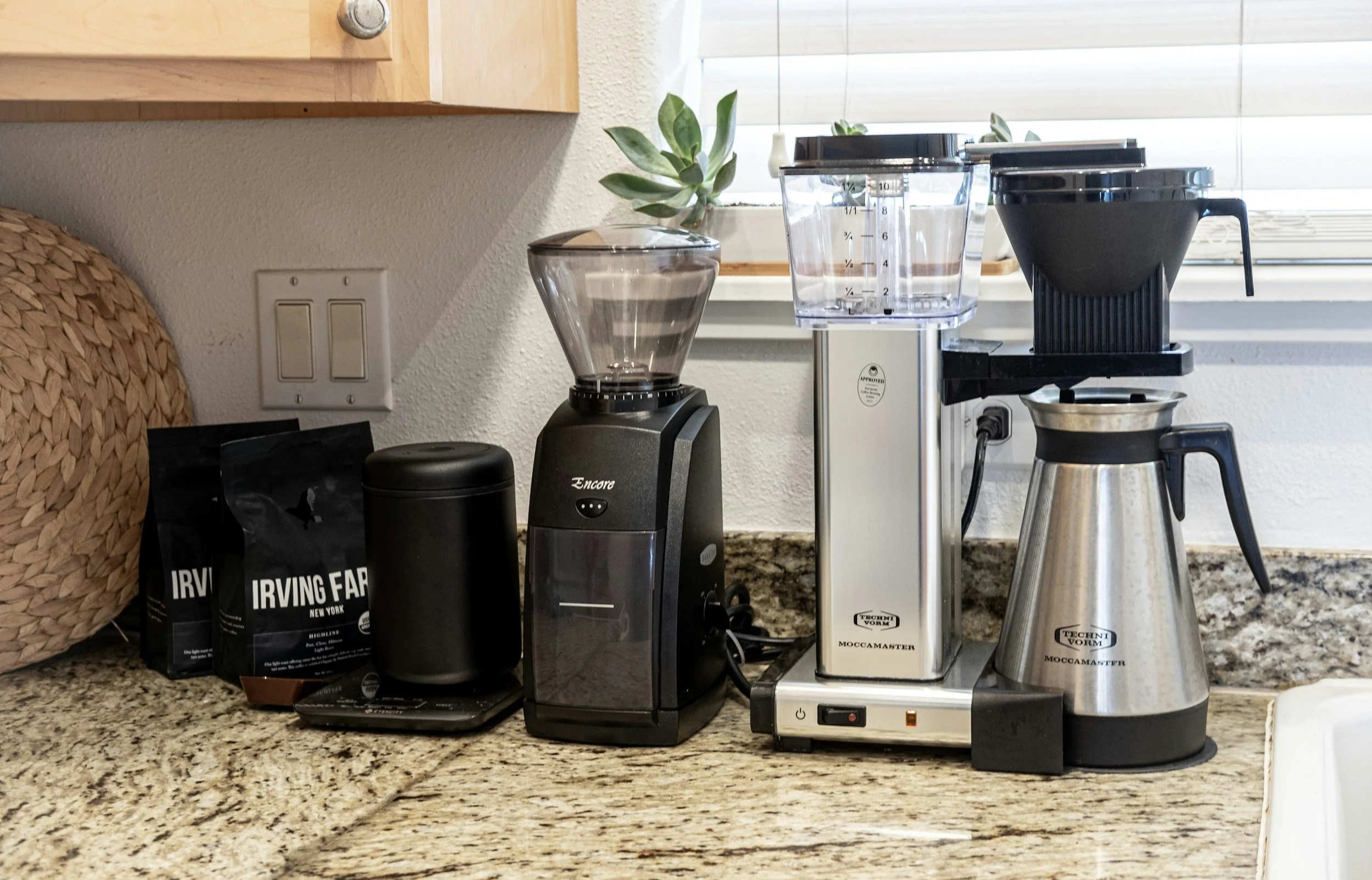 Coffee station with coffee bean bags, a black grinder, a water filter, a black pour-over coffee maker, and a stainless steel thermal carafe on a granite countertop.