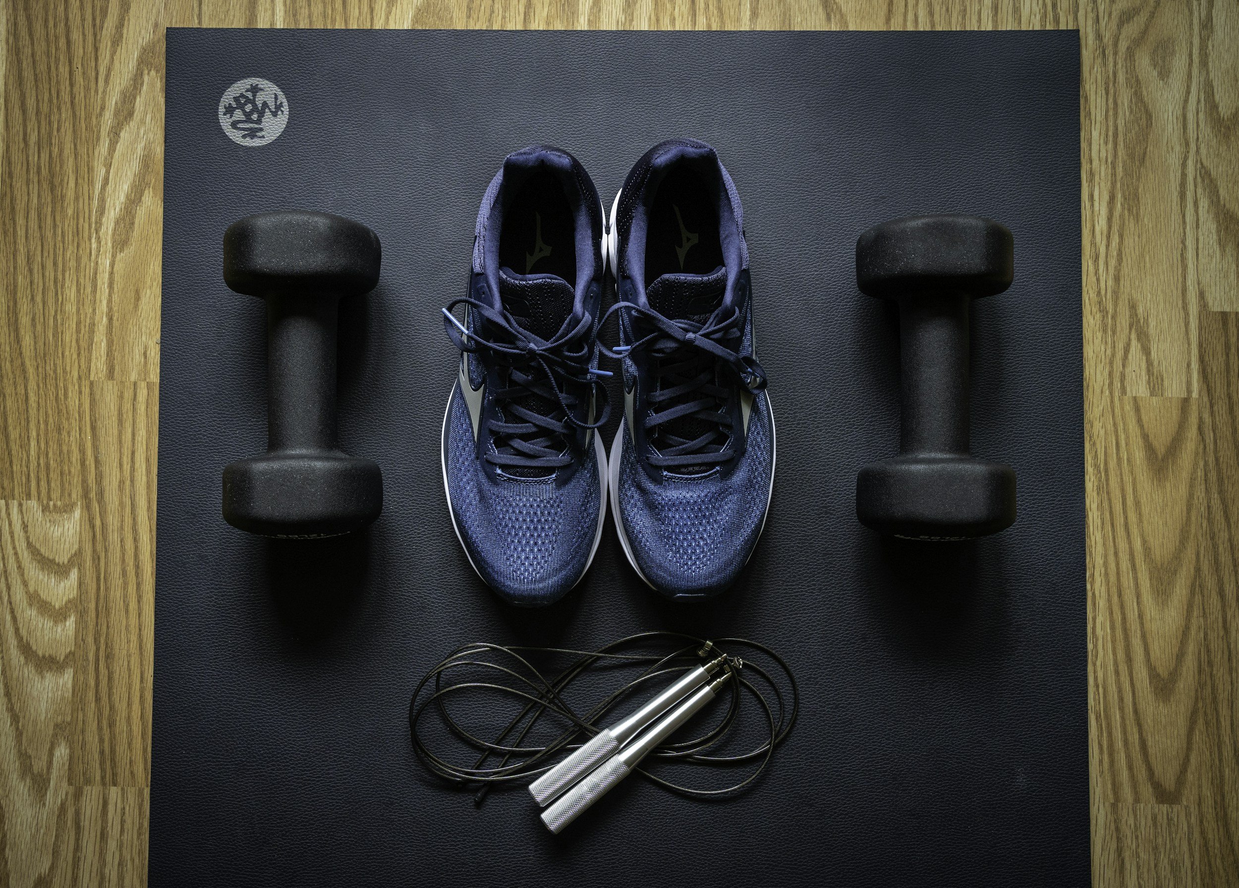 Pair of blue running shoes, two black dumbbells, and a jump rope placed on a black exercise mat on a wooden floor.