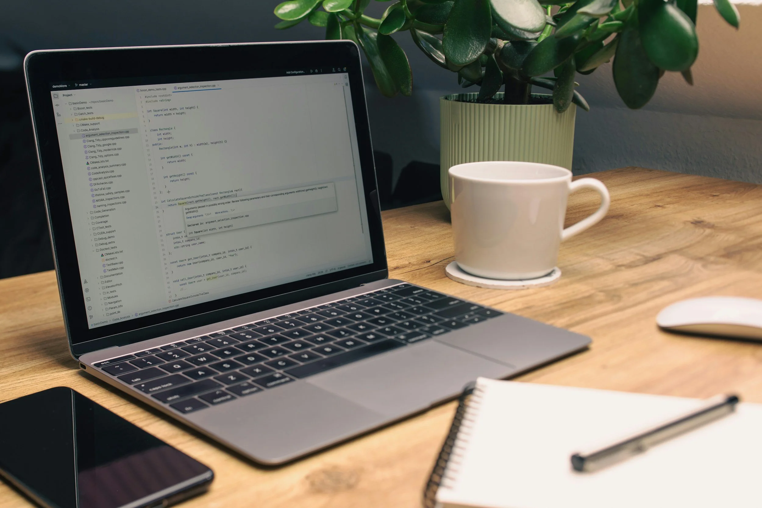 A laptop on a wooden desk displaying code, with a white coffee mug, a plant in a green pot, an iPhone, a white mouse, and a notebook with a pen, in a work or study space.