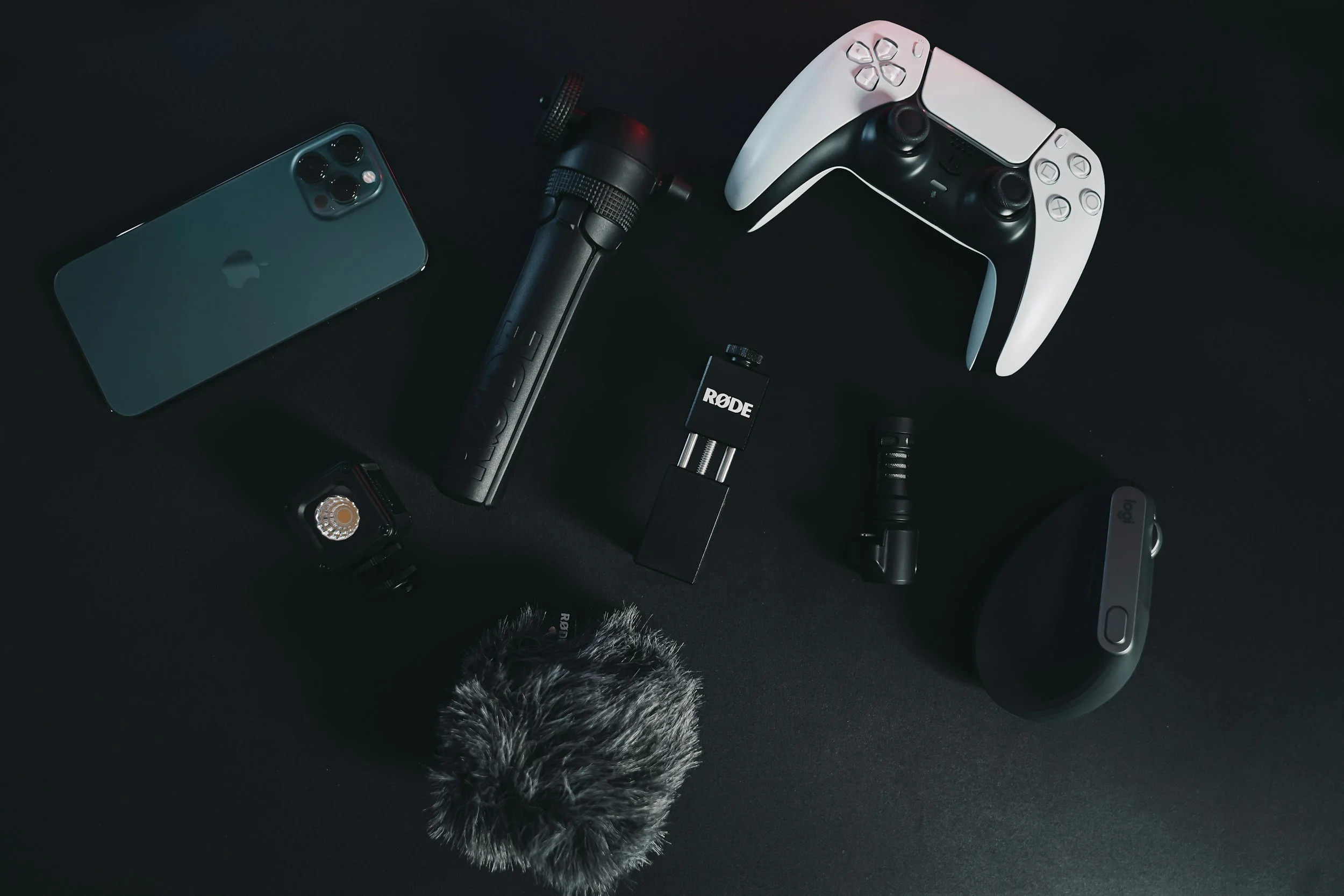 Various audio recording and gaming equipment including a smartphone, microphone, microphone windscreen, audio interface, handheld mic, gaming controller, and a computer mouse on a dark surface.