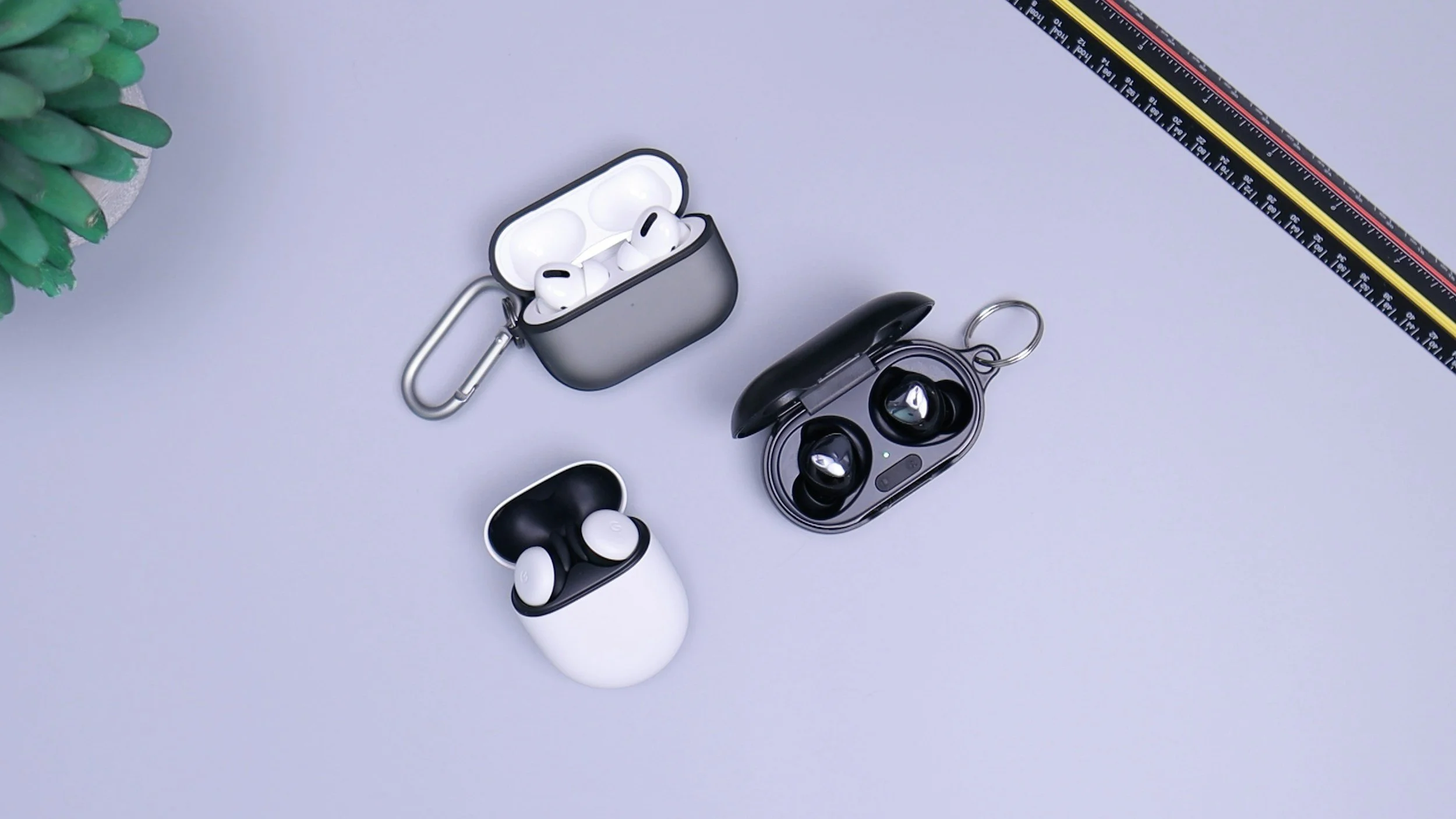 Wireless earbuds inside their cases, with a charging case and accessories on a light gray surface, with a ruler and a small green plant partially visible.