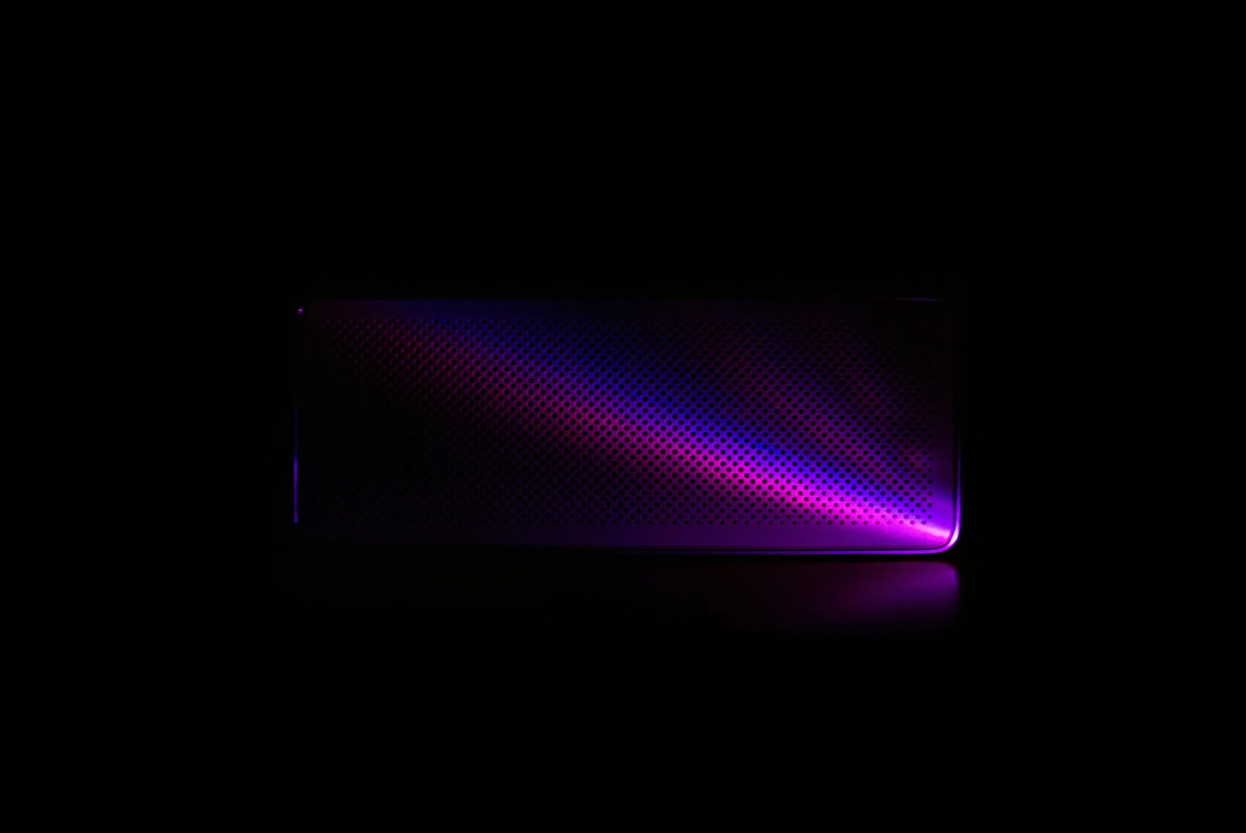 A dark, rectangular electronic device with a perforated surface, illuminated by purple and blue LED lights.