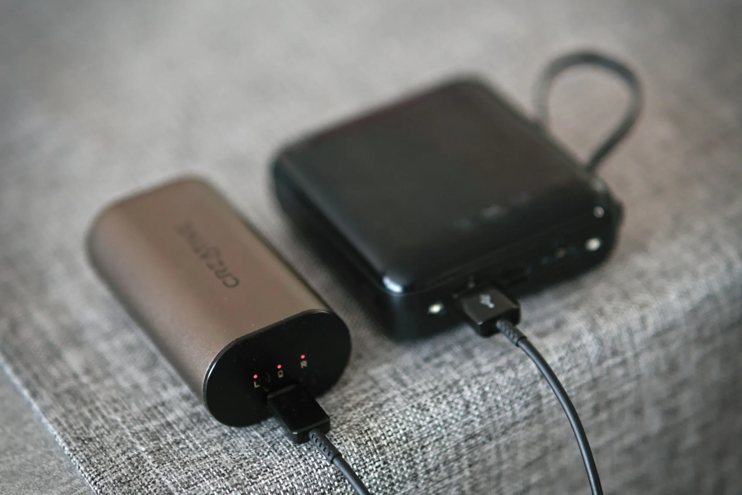 A charging dock connected to a power bank and a portable Bluetooth speaker, both placed on a gray fabric surface.
