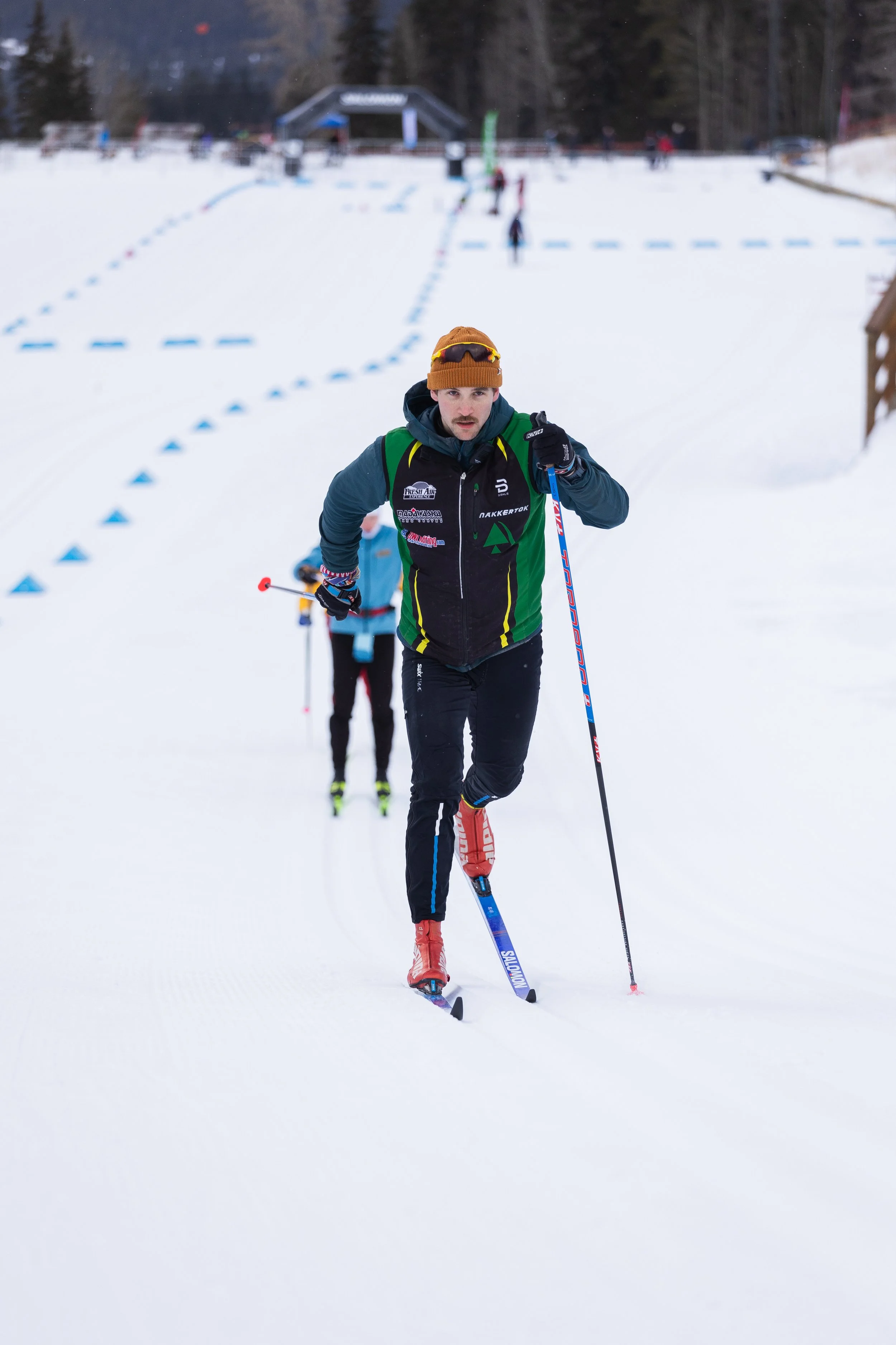 A man cross-country skiing on a snowy trail with trees in the background and other skiers in the distance.