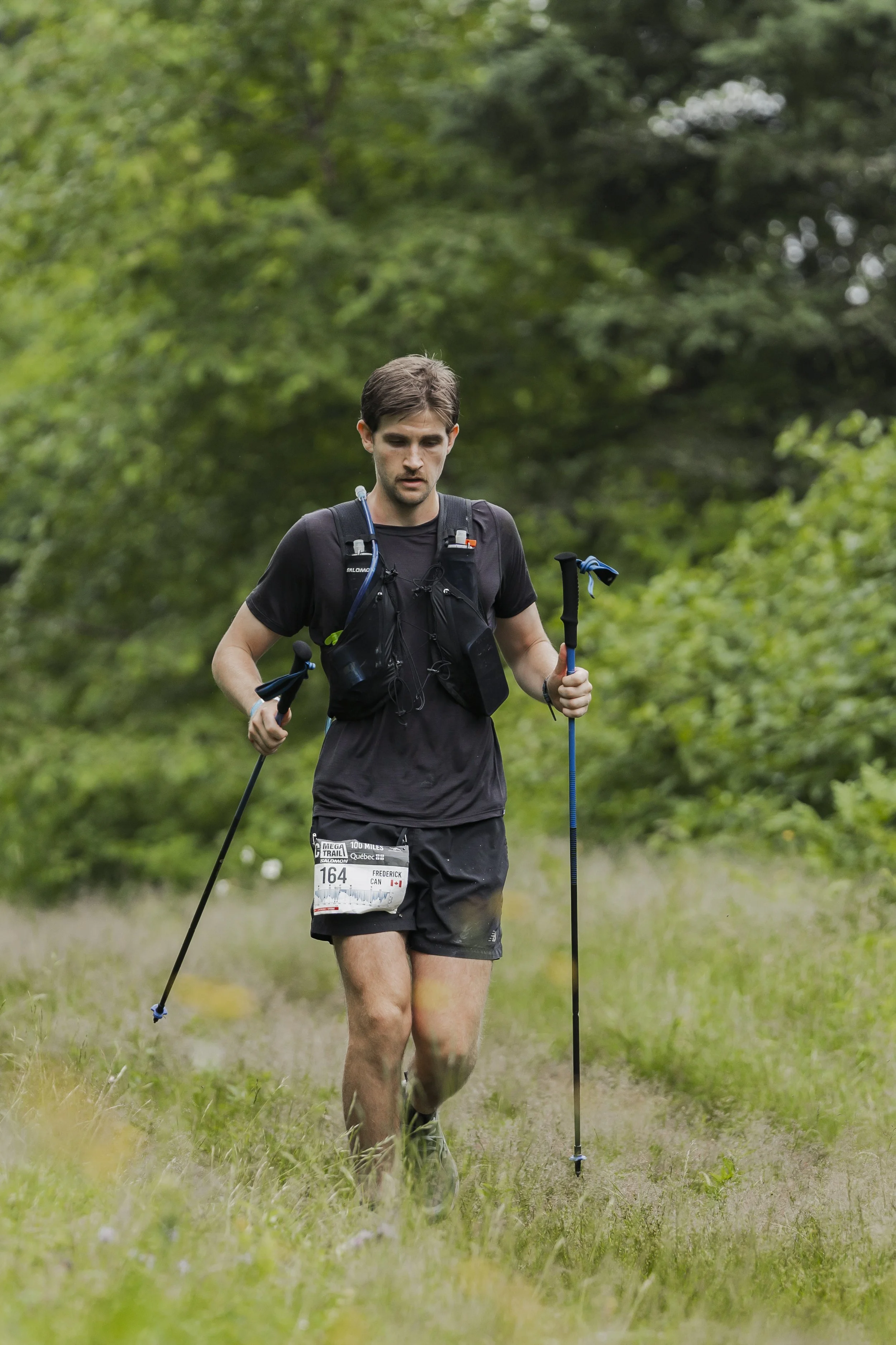 An athletic male runner in black running gear, carrying trekking poles and wearing a trail race bib, running through a grassy, wooded trail.