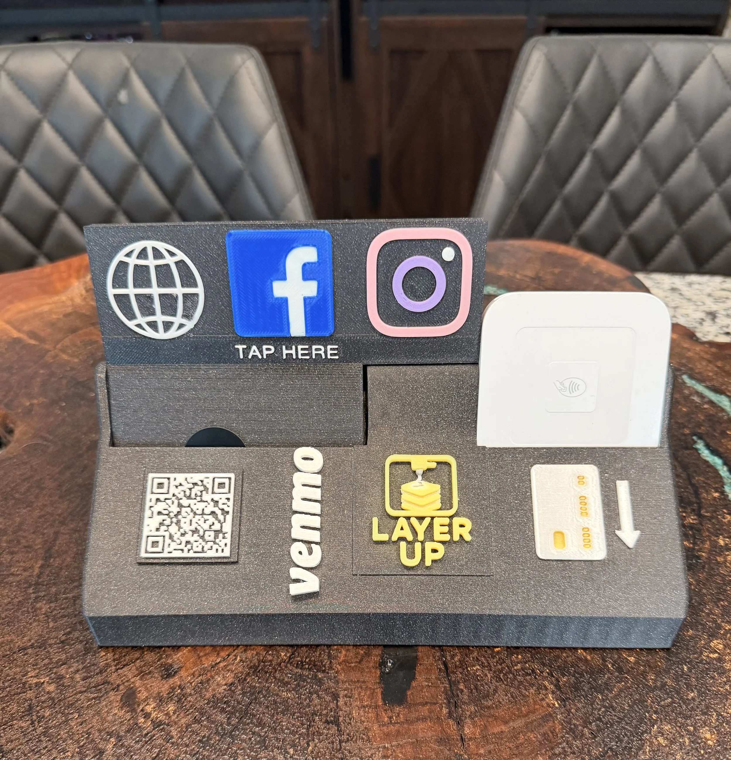 Close-up of social media and website icons on a board on a wooden table, with a QR code and QR code icon nearby, and chairs and wooden furniture in the background.