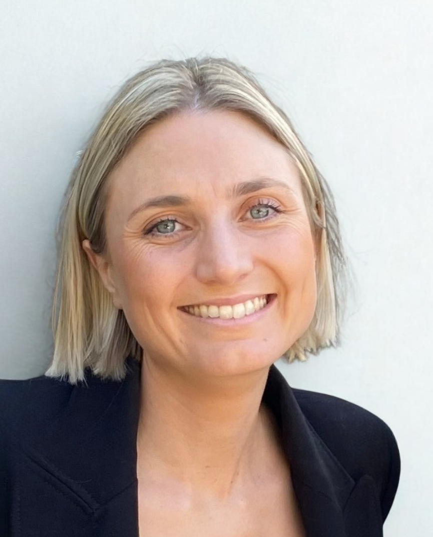 Close-up portrait of a smiling woman with blonde hair, blue eyes, and a black blazer, standing against a plain white background.
