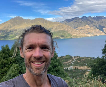 Man smiling outdoors with mountains and lake in the background.