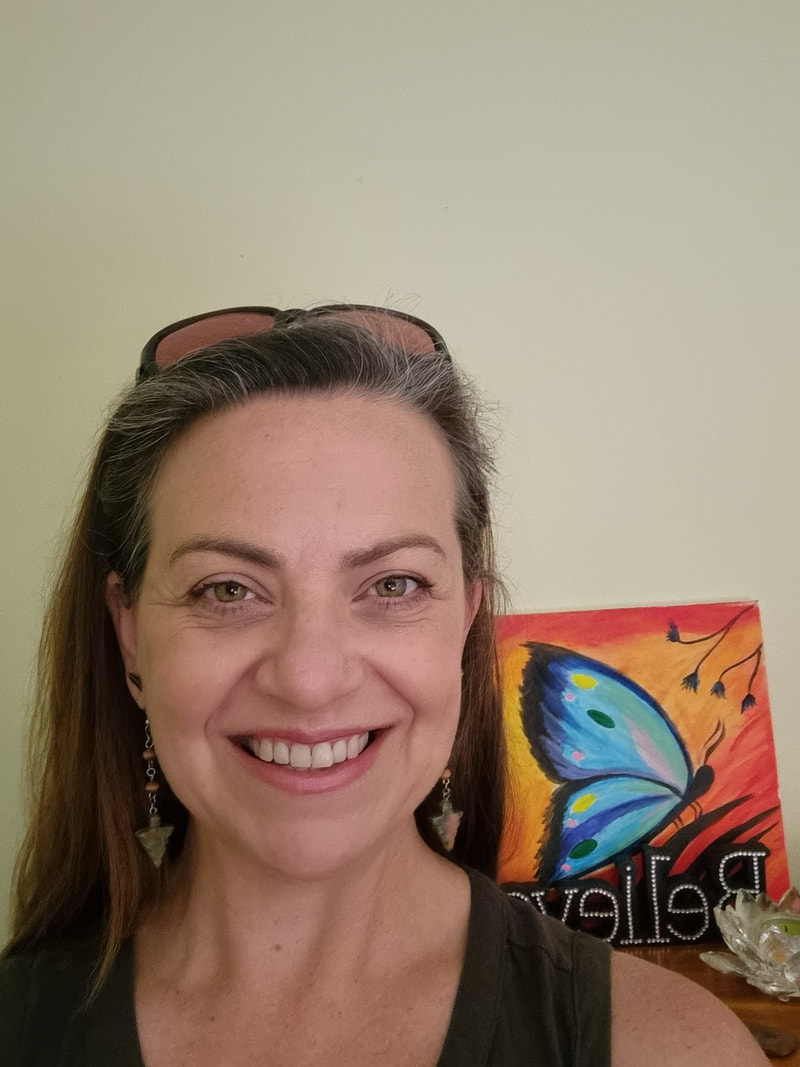 Smiling woman with long brown hair wearing earrings and sunglasses on her head, standing in front of a colorful butterfly painting and a decorative animal-shaped bowl on a table.