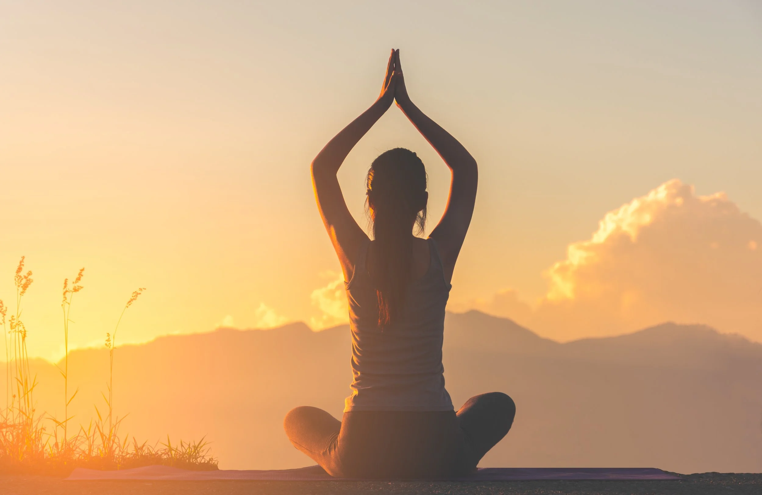 A person practicing yoga outdoors at sunrise, seated in a cross-legged position with hands pressed together overhead.