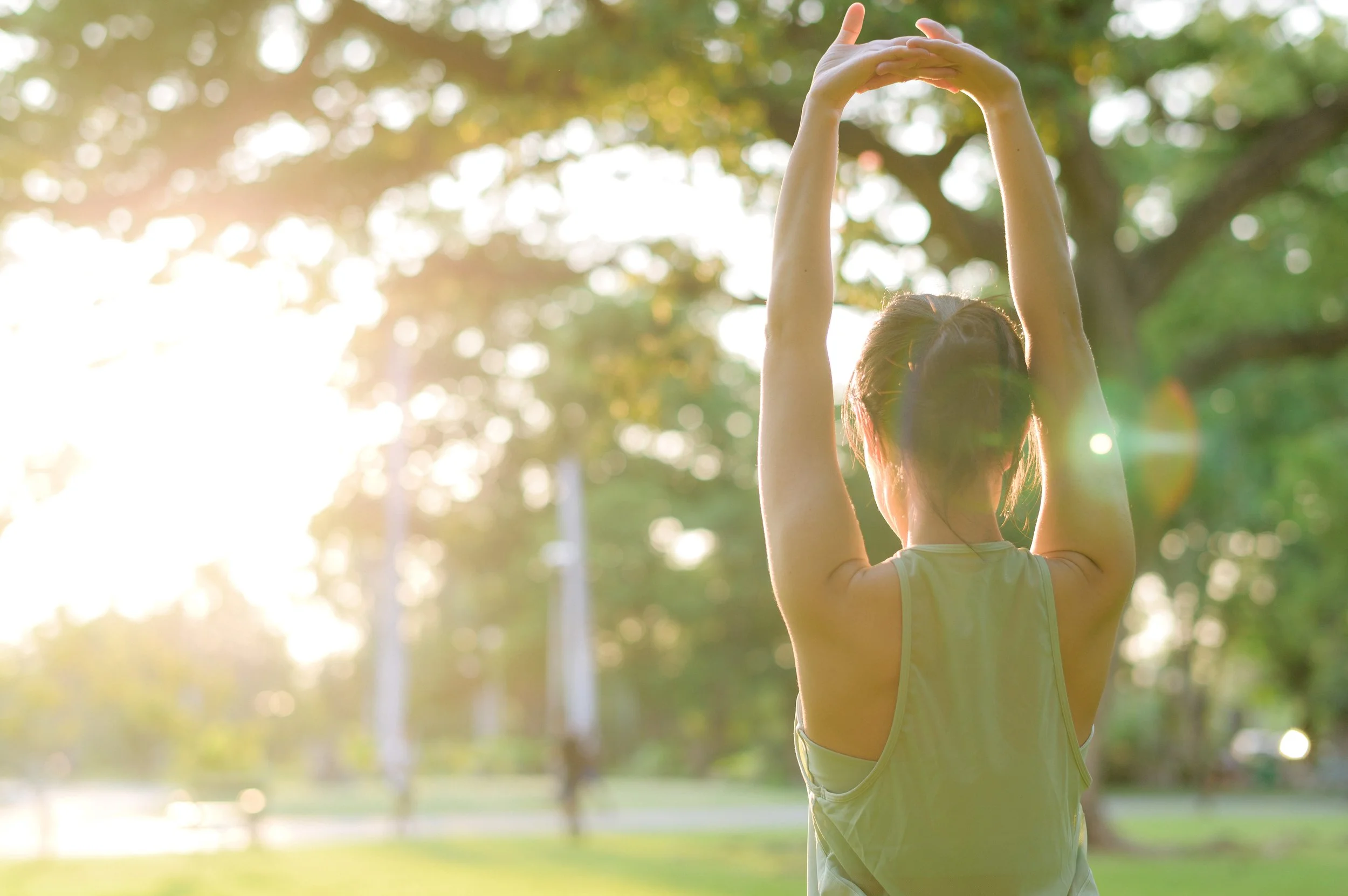 A woman in a yellow tank top standing outdoors in a park, stretching with her arms raised above her head during sunset, with sunlight filtering through the trees.