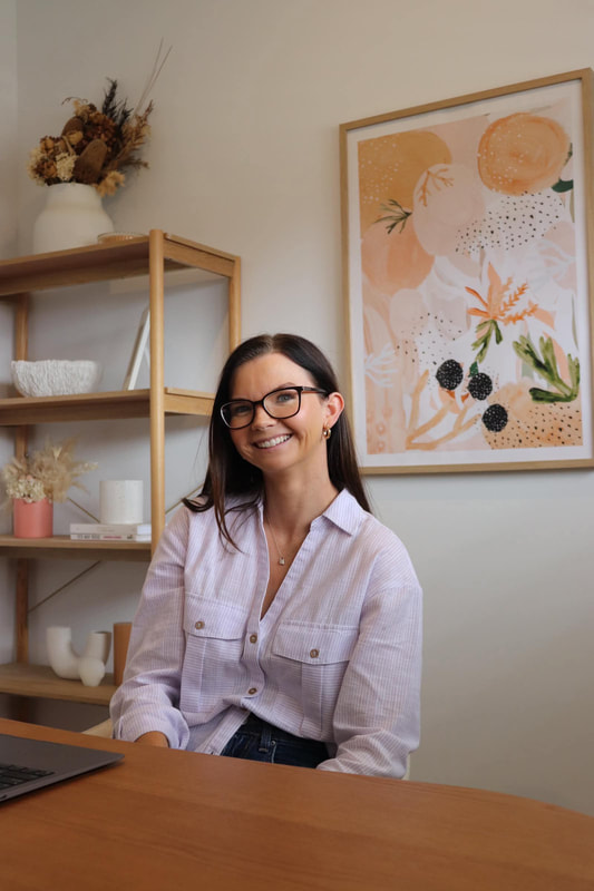 A woman with glasses smiling while sitting at a wooden desk in a decorated room with shelves and an abstract art piece on the wall.