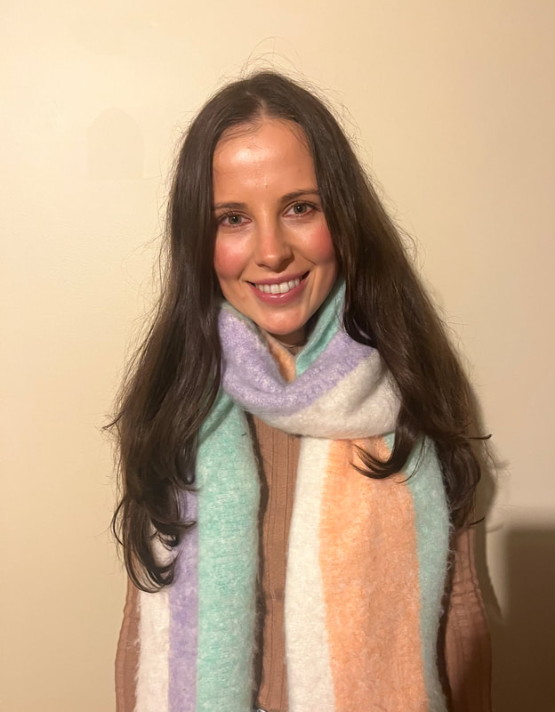 A young woman with long brown hair is smiling at the camera, wearing a colorful striped scarf and a brown top, standing against a beige wall.