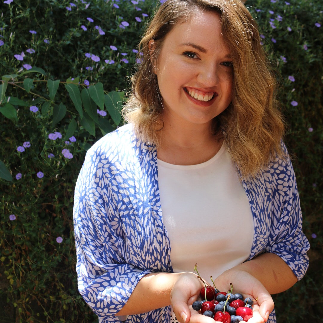 A smiling woman holding a handful of cherries, standing outdoors in front of green foliage and purple flowers.
