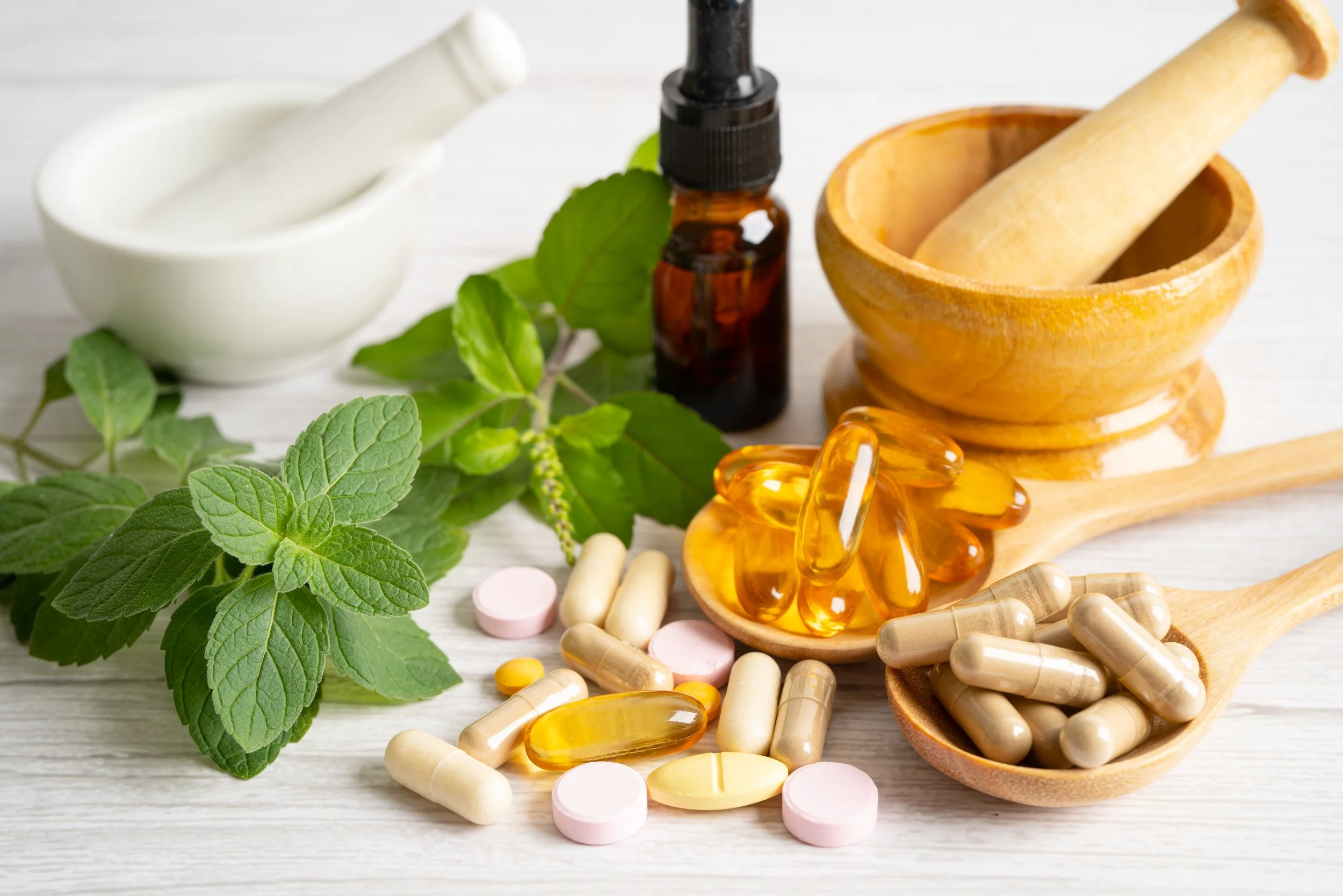 Fresh herbal mint leaves, various pills and capsules, a brown herbal extract bottle, a mortar and pestle, and a wooden spoon with gel capsules on a white wooden surface.