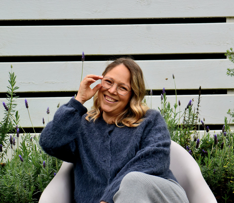 A smiling woman with glasses sitting outdoors in front of a white fence with purple flowers, wearing a cozy navy sweater.