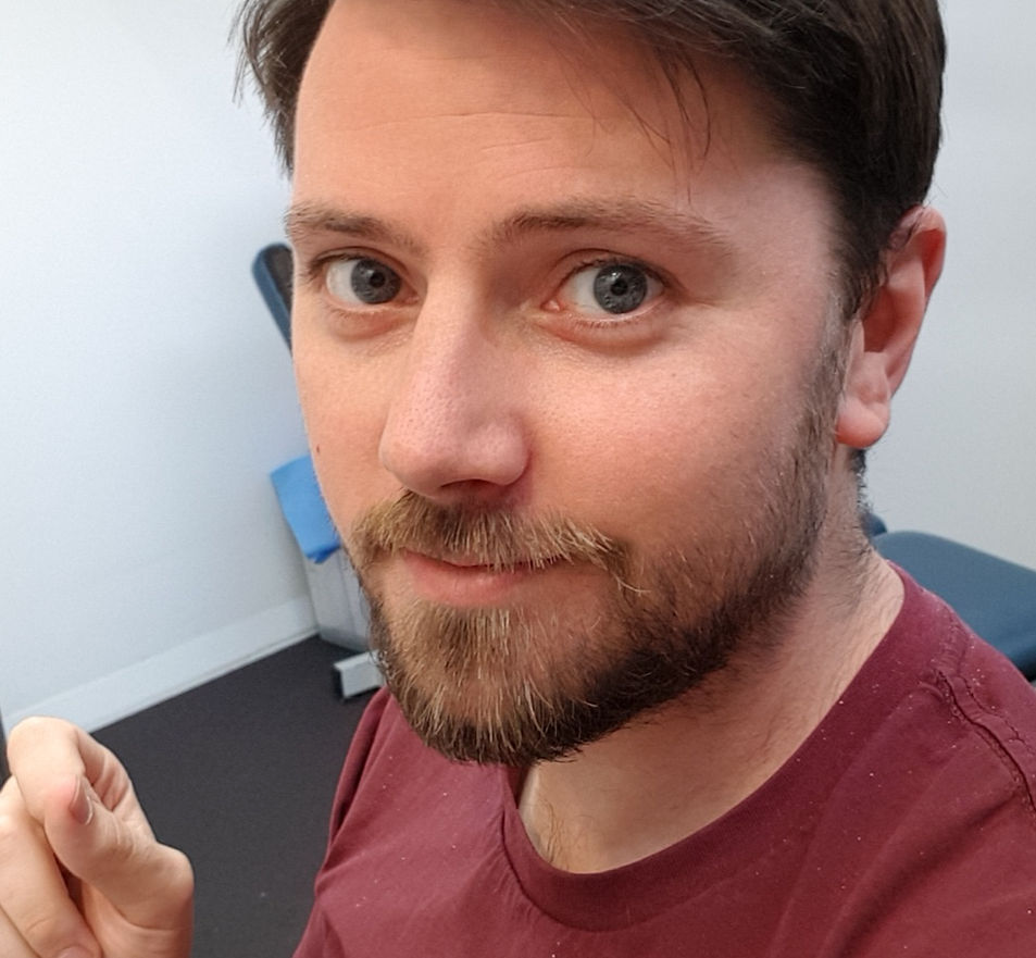 A man with light brown hair, a beard, and blue eyes taking a selfie indoors, wearing a maroon shirt.