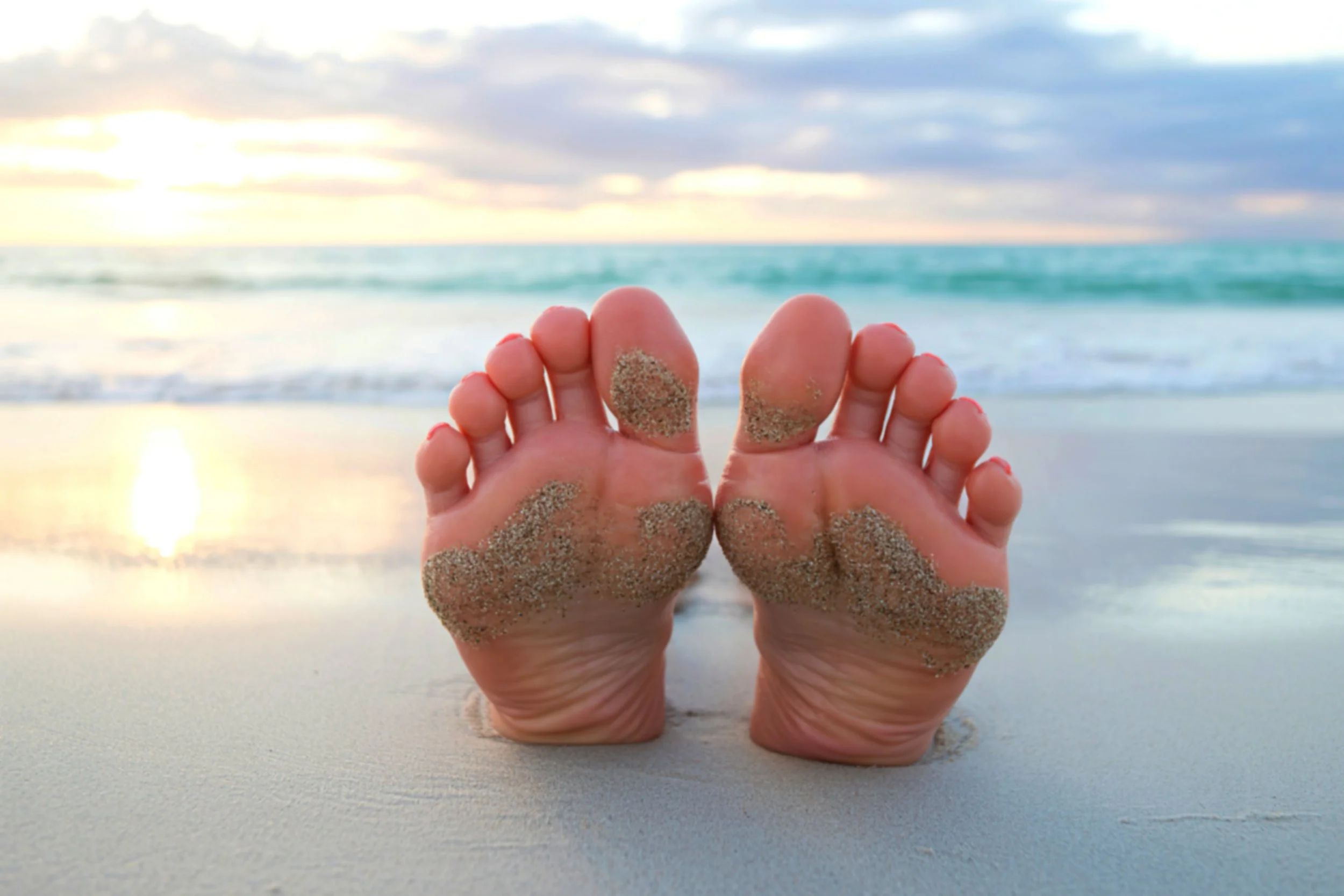Close-up of sandy feet on a beach with the sunrise over the ocean in the background.