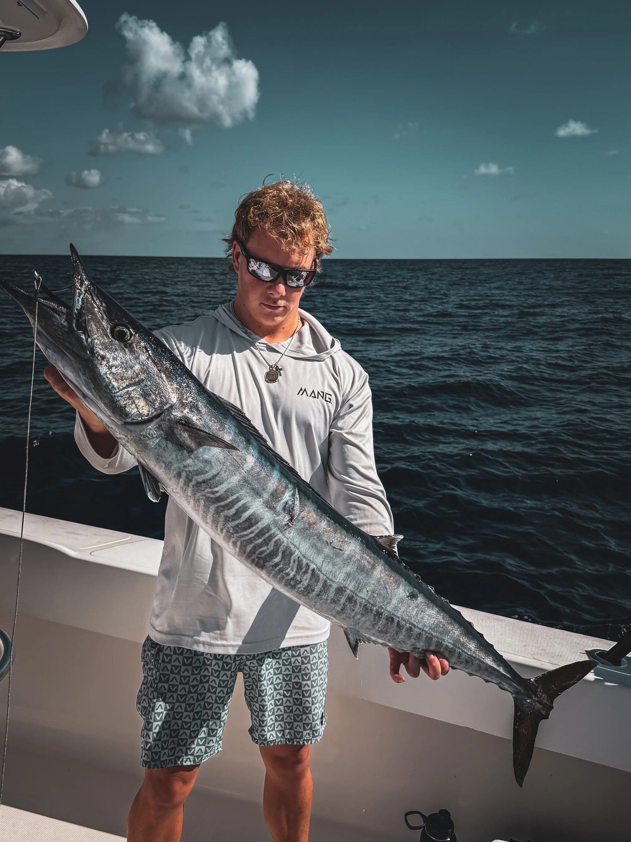 A young man wearing sunglasses and a white hoodie holding a large fish on a boat in the ocean under a partly cloudy sky.
