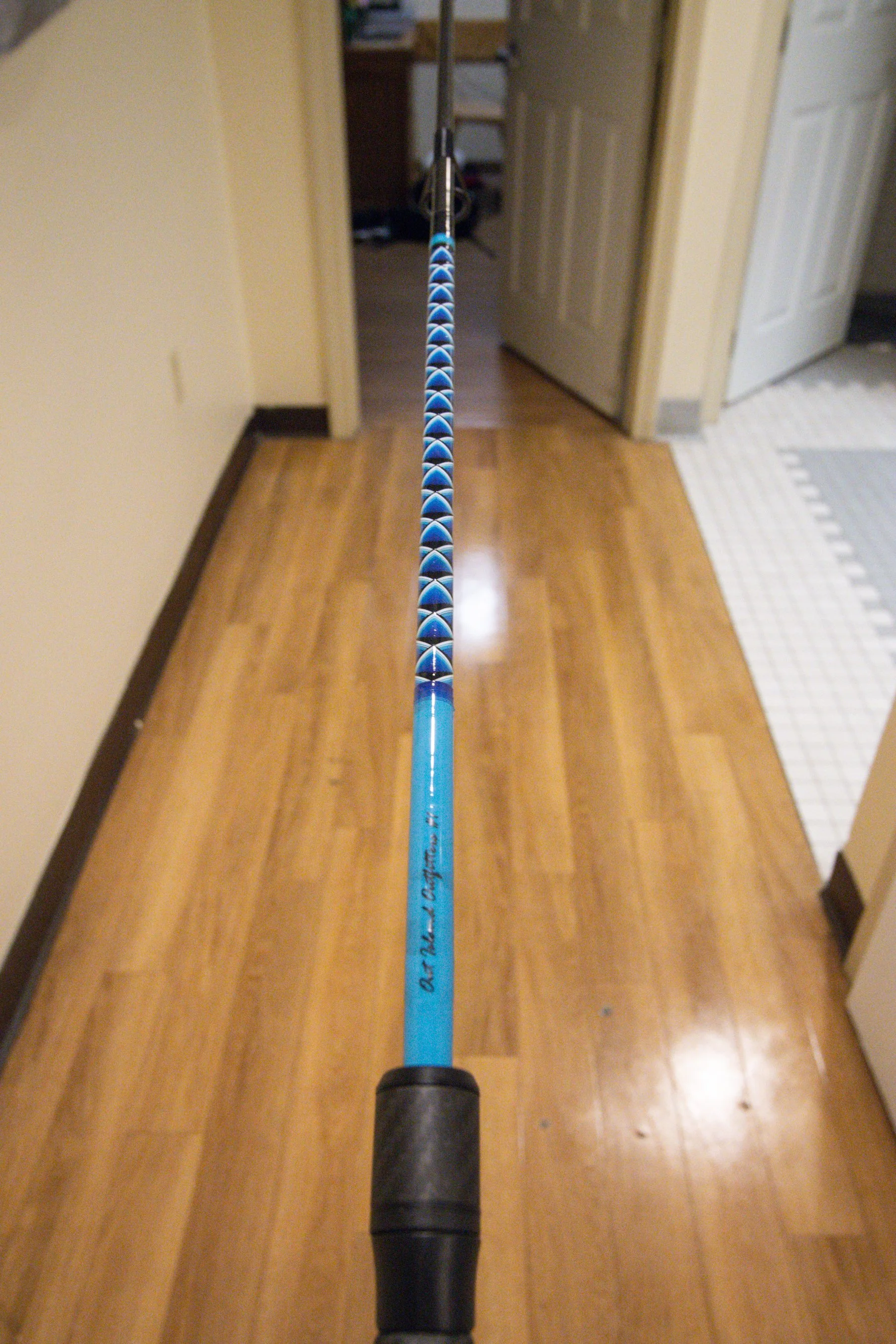 A fishing rod extended into a room with wooden floors, viewed from the handle end towards the room’s open doorway.