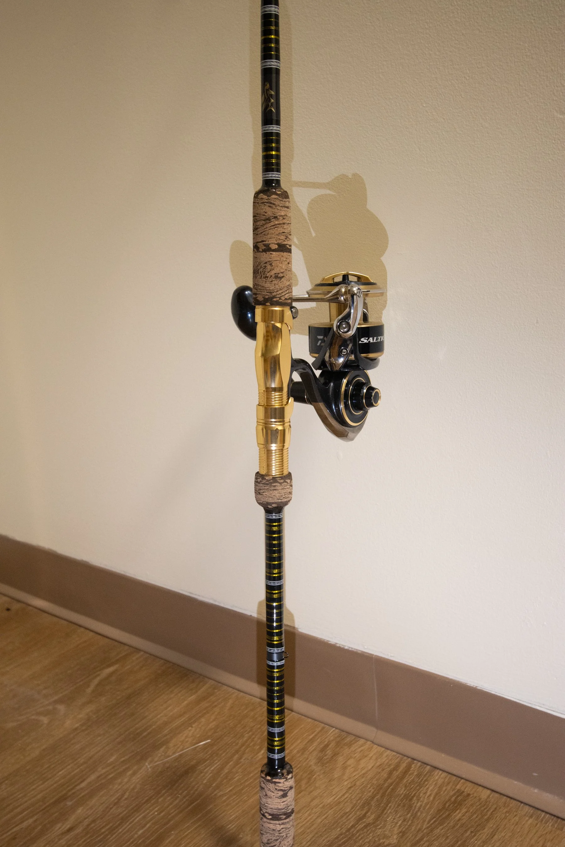 A fishing rod with a black and gold reel leaning against a beige wall on a wooden floor.
