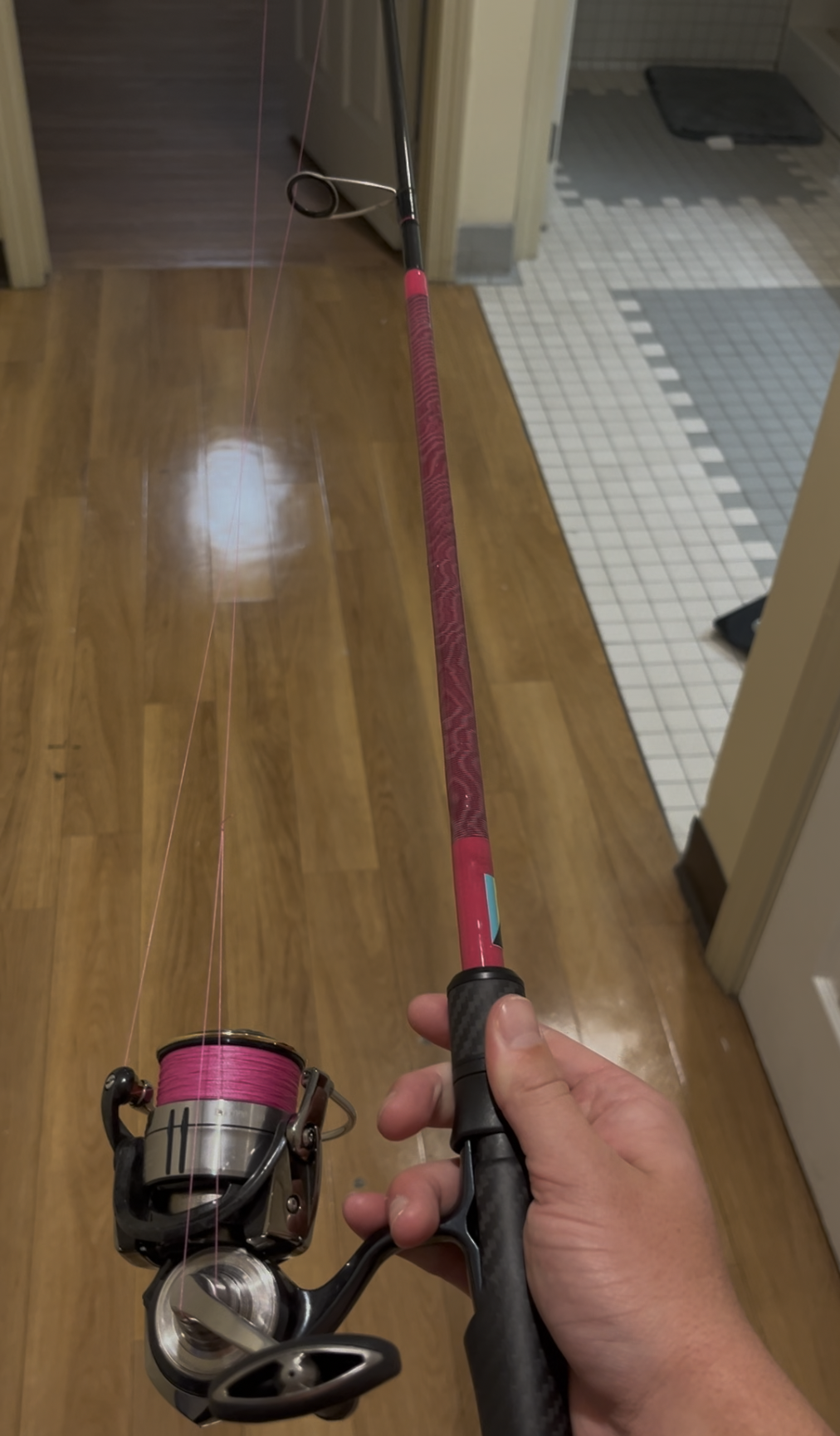A person holding a pink fishing rod with a spinning reel with pink fishing line, indoors on a wooden floor near a doorway.