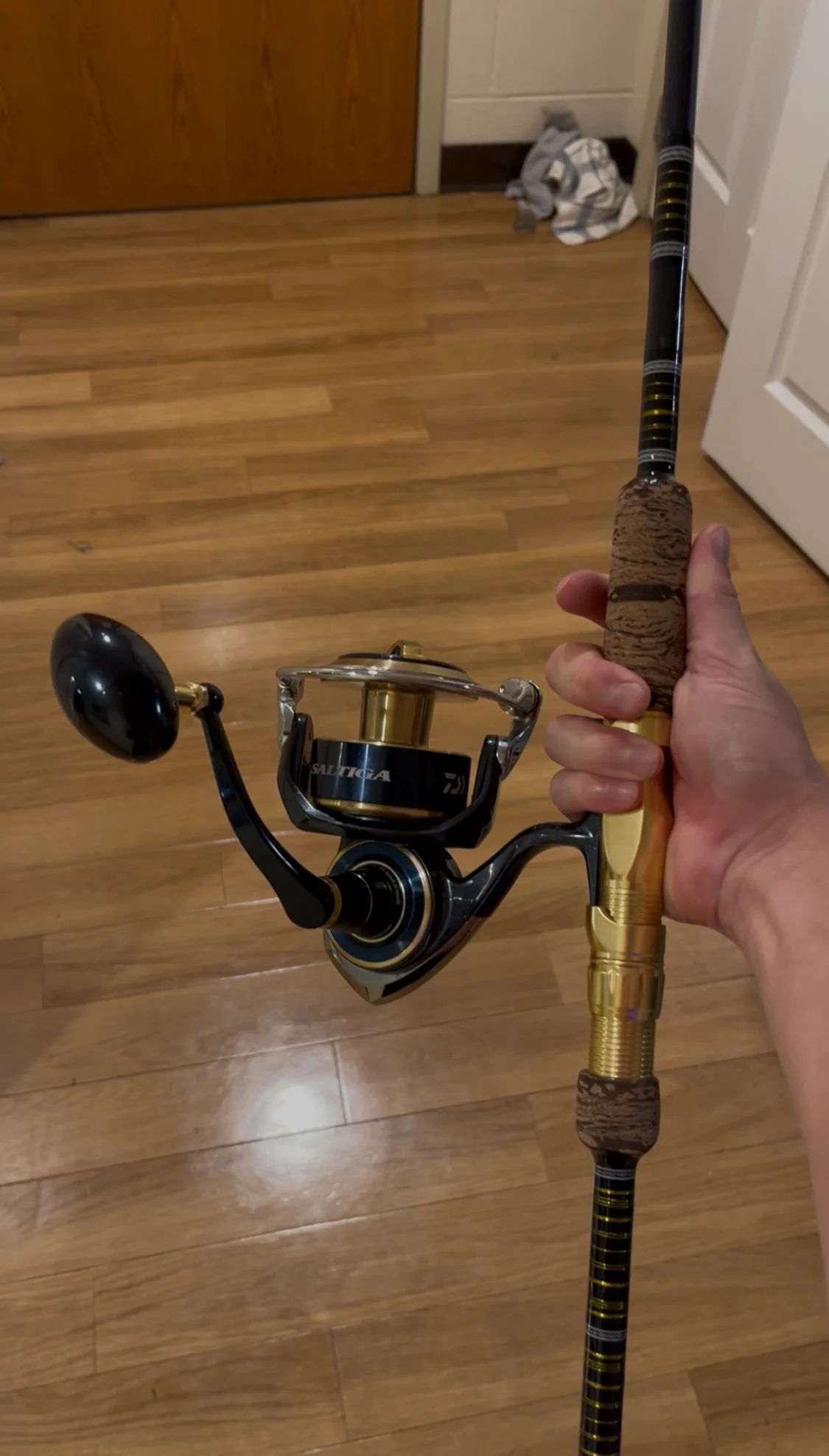 Hand holding a fishing rod with a reel, indoors on wooden floor.