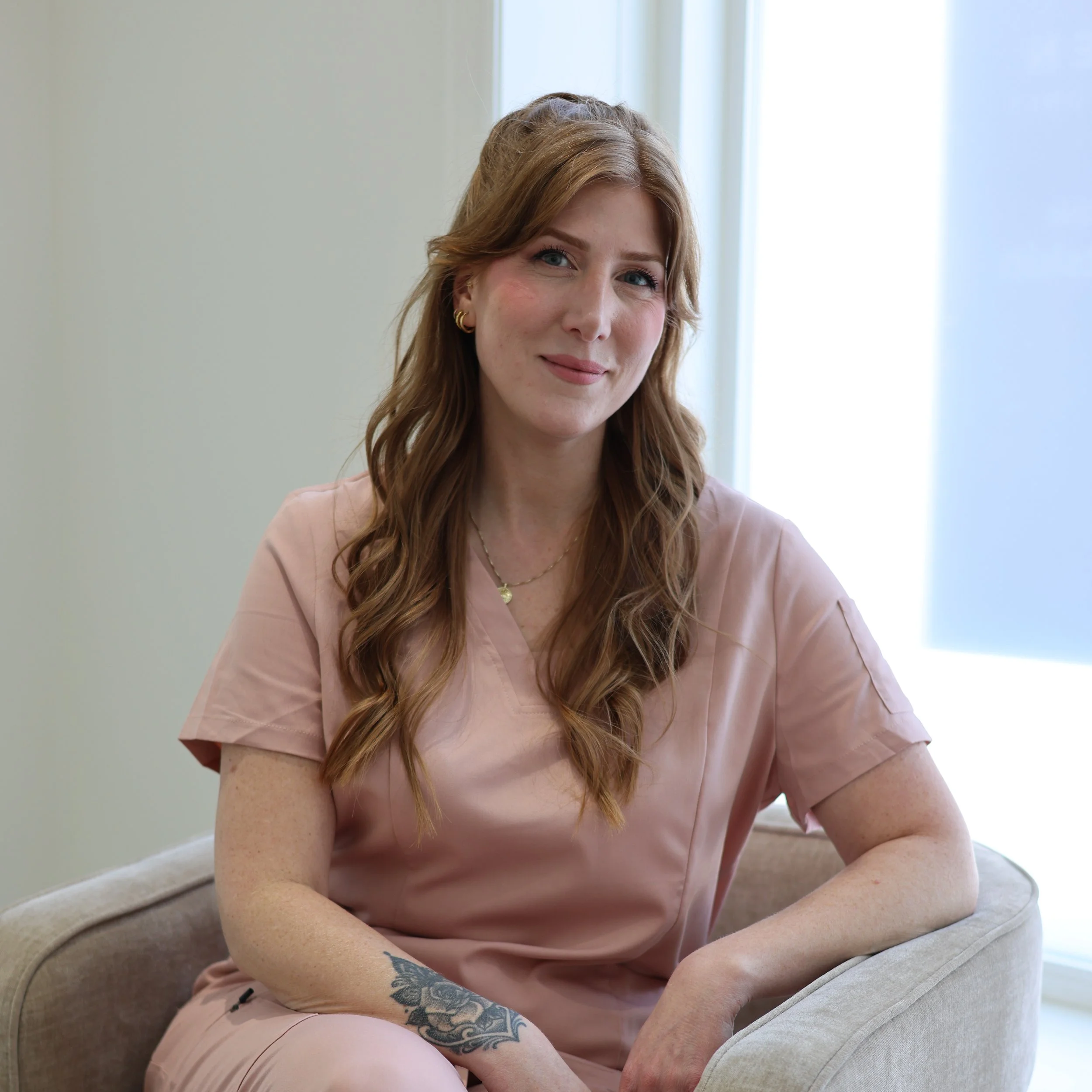 A young woman with long red hair, blue eyes, and light skin, smiling softly, sitting on the floor near a window with natural light, wearing a pink t-shirt and earrings, with a tattoo of flowers on her forearm.