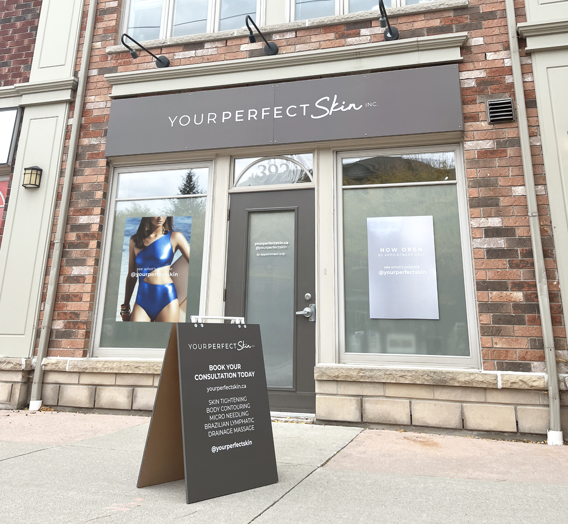Exterior view of a storefront with a grey sign reading 'YOURPERFECTSkin INC.' above the entrance, located at house number 392. A black sandwich board sign on the sidewalk advertises skin treatments and book appointment. The storefront has large windows, and outside promotional posters are displayed inside the windows. The building features brick walls and decorative trim.