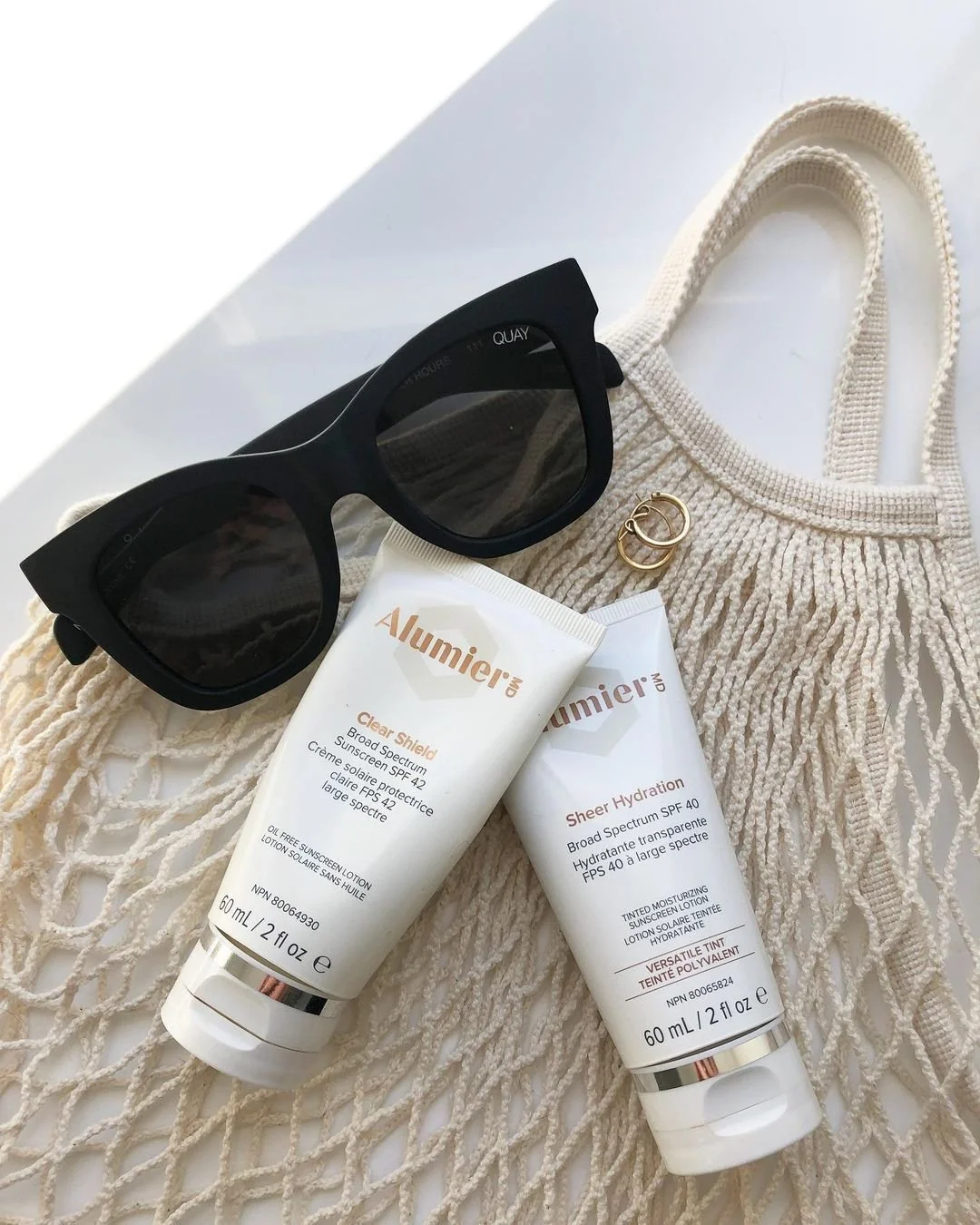 A flat lay of a white knitted tote bag with woven handles, black sunglasses, a pair of gold hoop earrings, and two tubes of sunscreen lotion on a white surface.