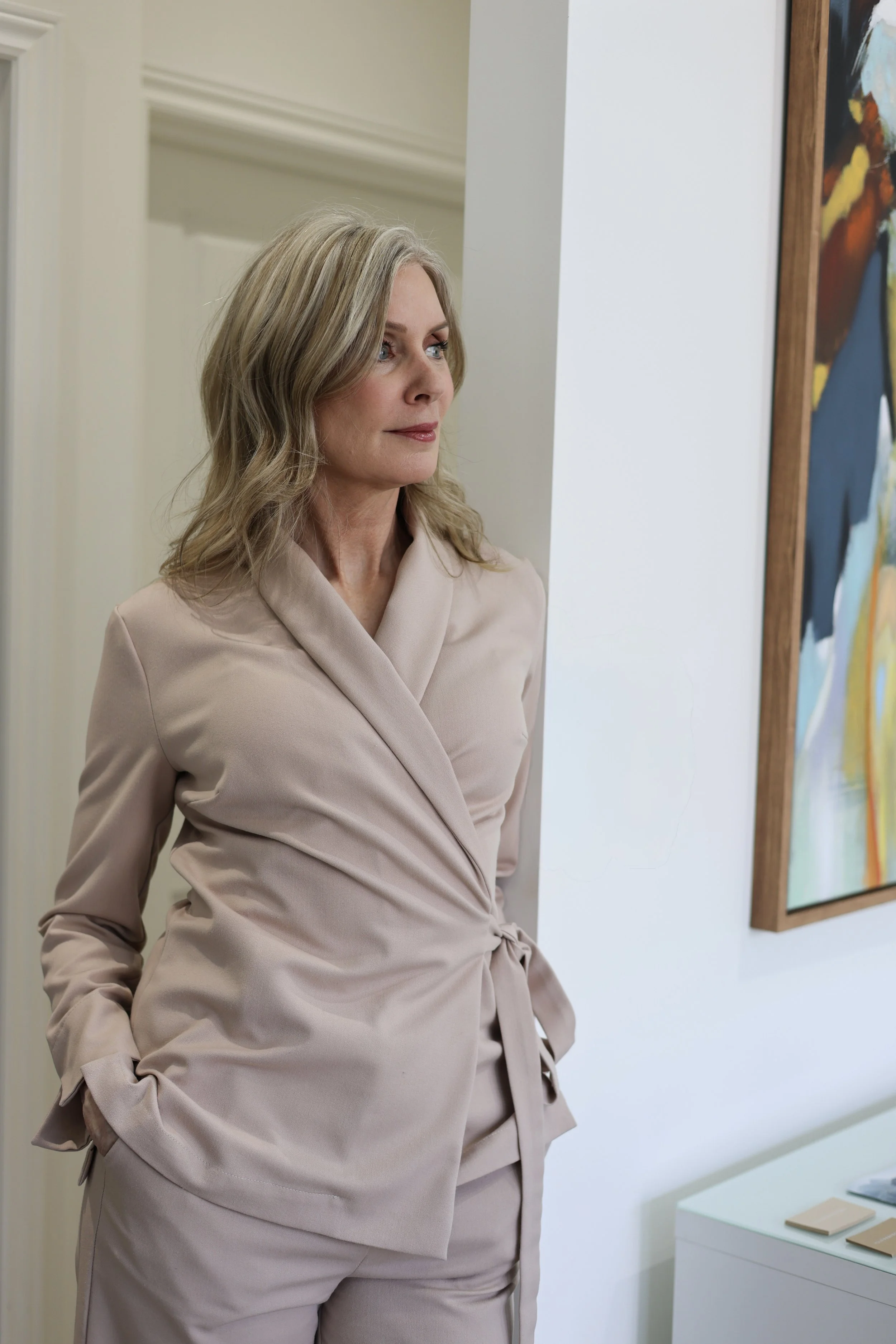 A woman with blonde hair wearing a beige blazer and matching pants, standing and looking at a colorful abstract painting in an art gallery or office setting.